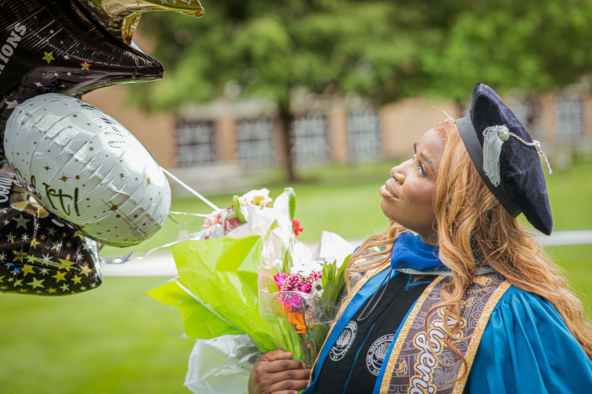 A young woman with long red hair wearing a graduation cap and gown standing outdoors, holding a bouquet of flowers, and looking at someone handing her balloons.