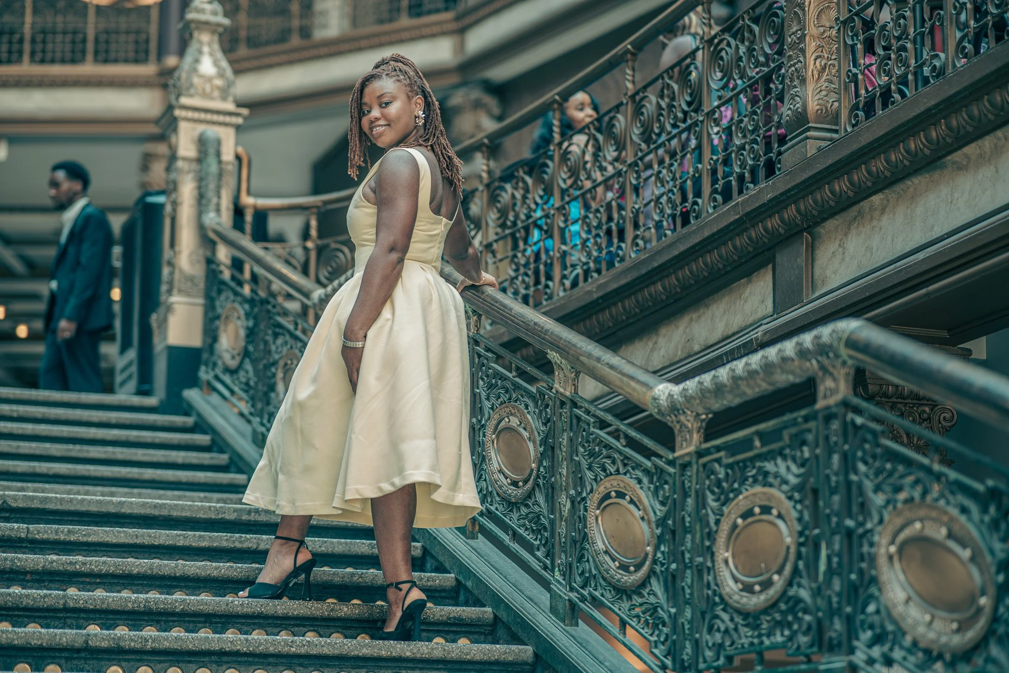 Woman in a beige satin dress and black high heels standing on a grand staircase with ornate wrought iron railing, smiling at camera, in an elegant indoor setting with other well-dressed people in the background.