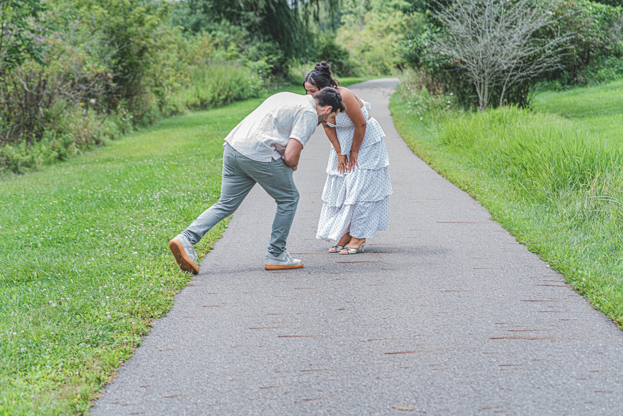 A man and woman standing on a paved path in a park, leaning toward each other and laughing under green trees.