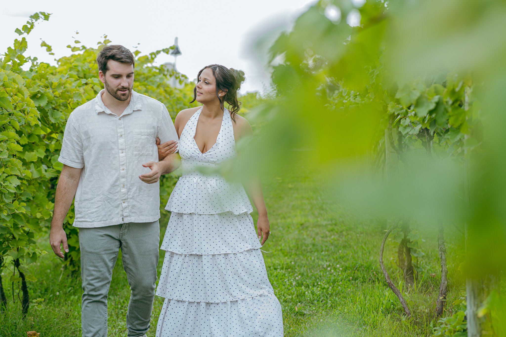 A man and a woman walking through a vineyard, with the woman touching the man's arm, surrounded by lush green grapevines