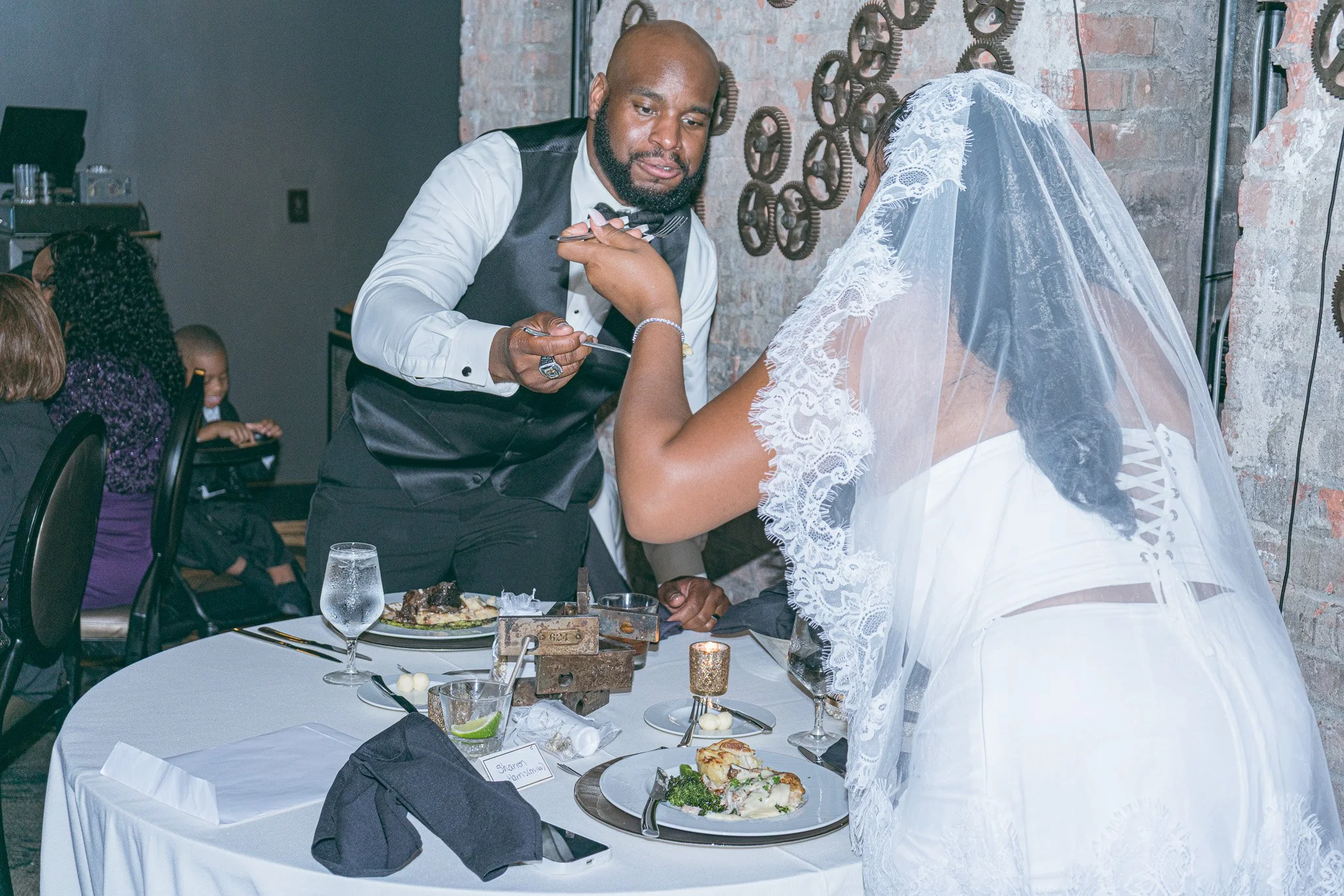 A man and a woman dressed in wedding attire sharing a moment during a wedding reception. The man is leaning forward towards the woman, who is wearing a lace wedding dress and veil. The reception table has plates of food, glasses, and decorative items.
