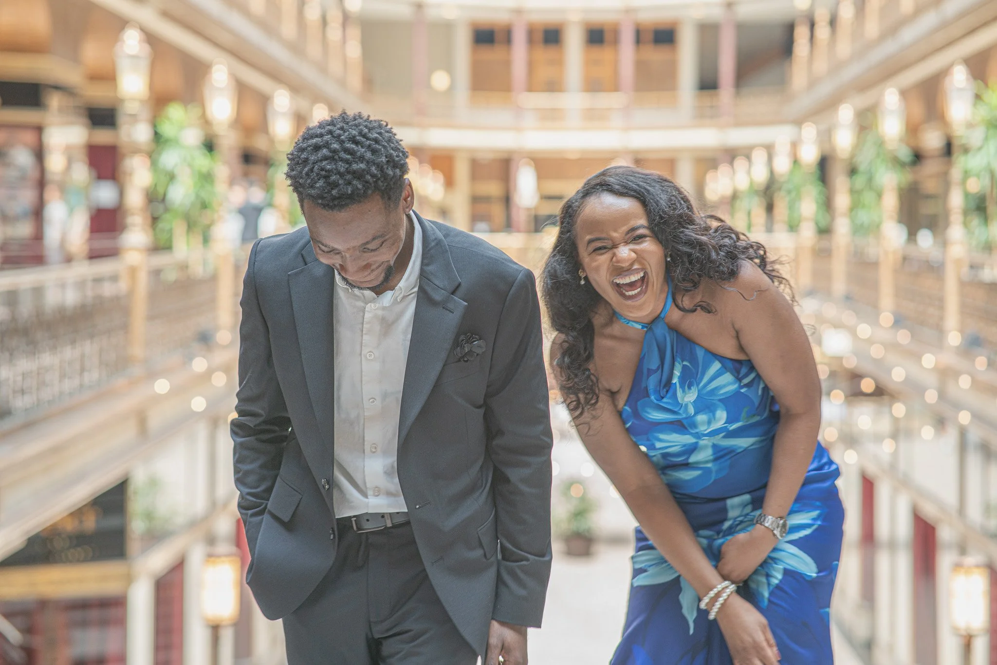 A man in a formal suit and a woman in a blue dress are laughing and bending forward, sharing a joyful moment in a brightly lit, elegant indoor setting with wooden balconies and greenery.