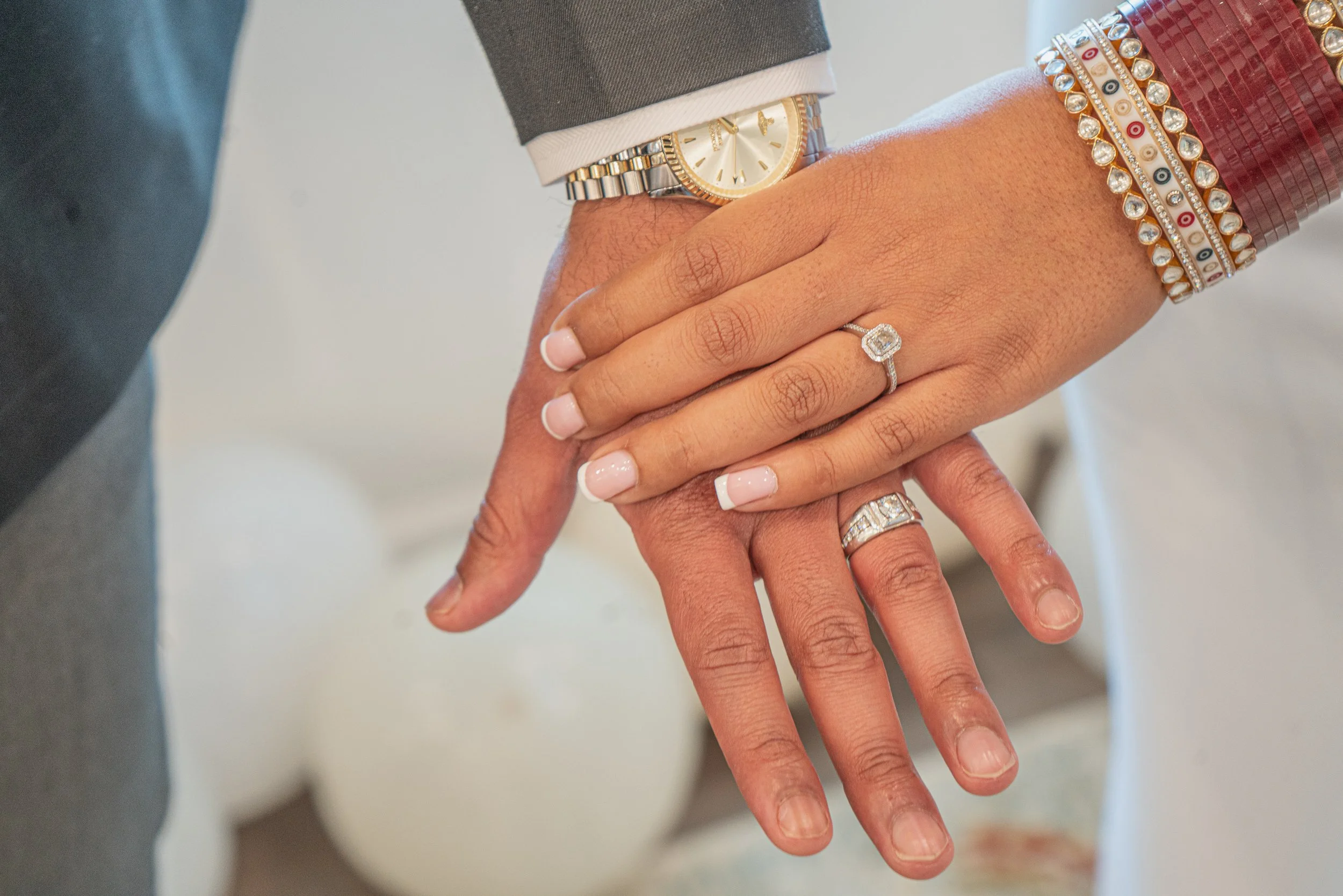 Close-up of the newlyweds two hands, one wearing a gray suit, both with wedding rings, a men's watch, and the bride wearing multiple bracelets as they hand smoothly lay over one another.