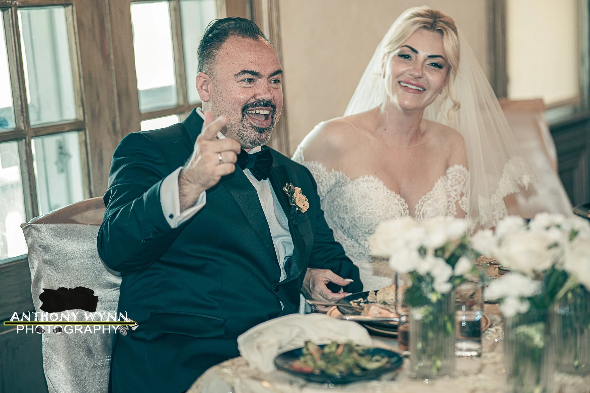 A man in a tuxedo and a woman in a wedding dress are sitting at a table during a wedding reception. The man is smiling and pointing, and the woman is smiling with a veil on her head.