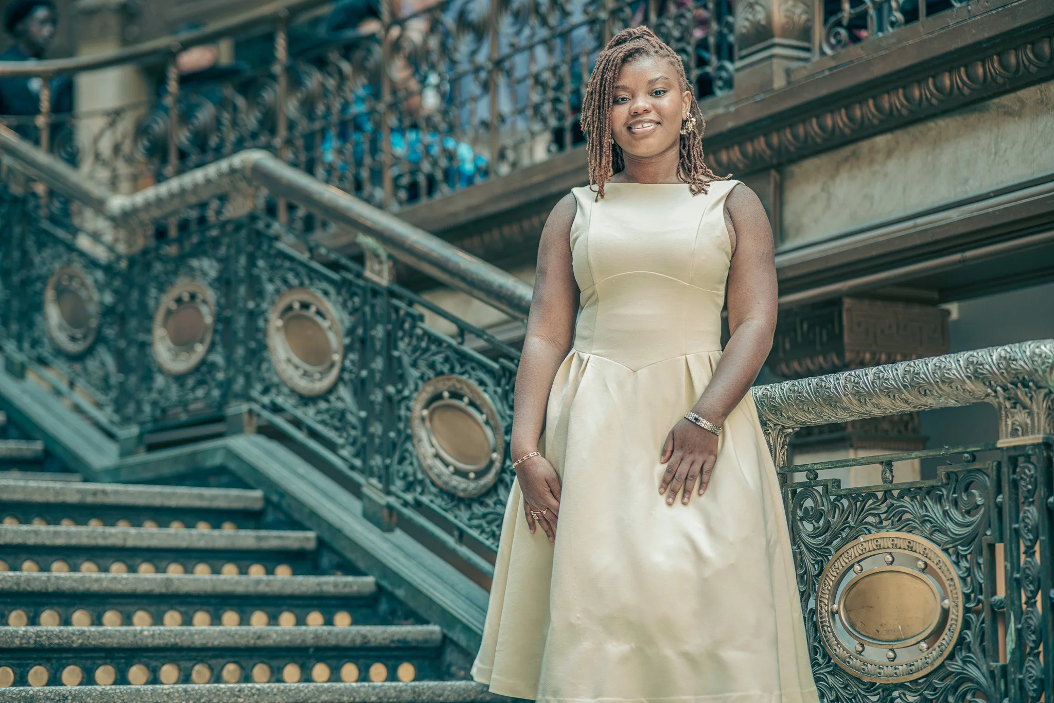 A woman in a cream-colored dress with a fitted bodice and full skirt, standing on an ornate staircase with decorative black wrought iron railings.