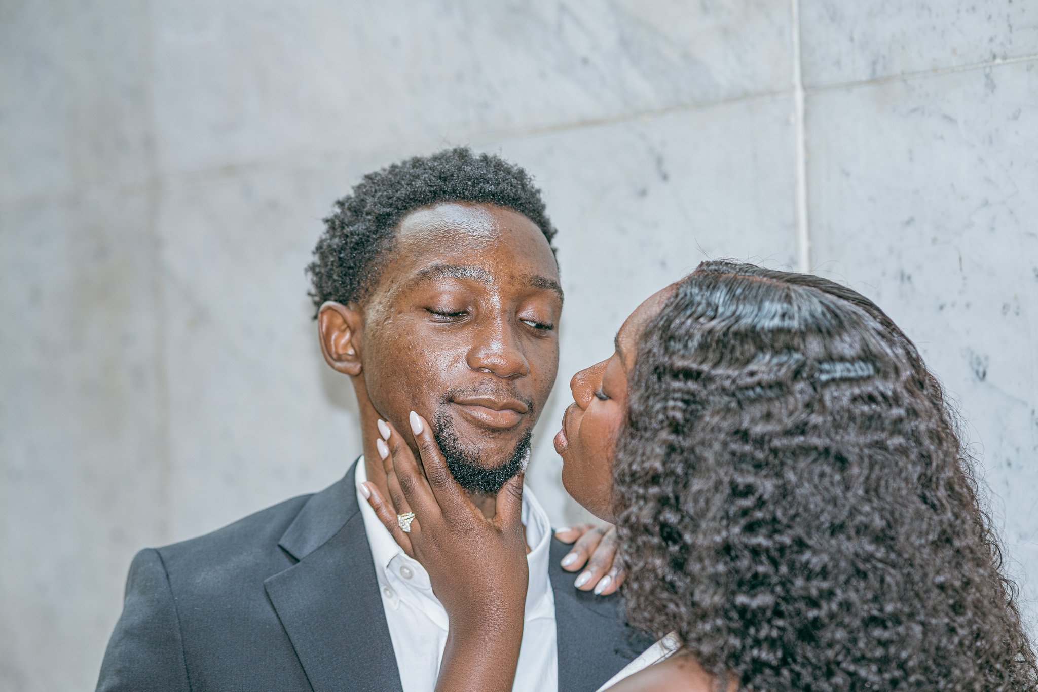 A woman and man couple in wedding attire in an intimate moment, with the woman touching the man's face and both with their eyes closed against a plain wall background.