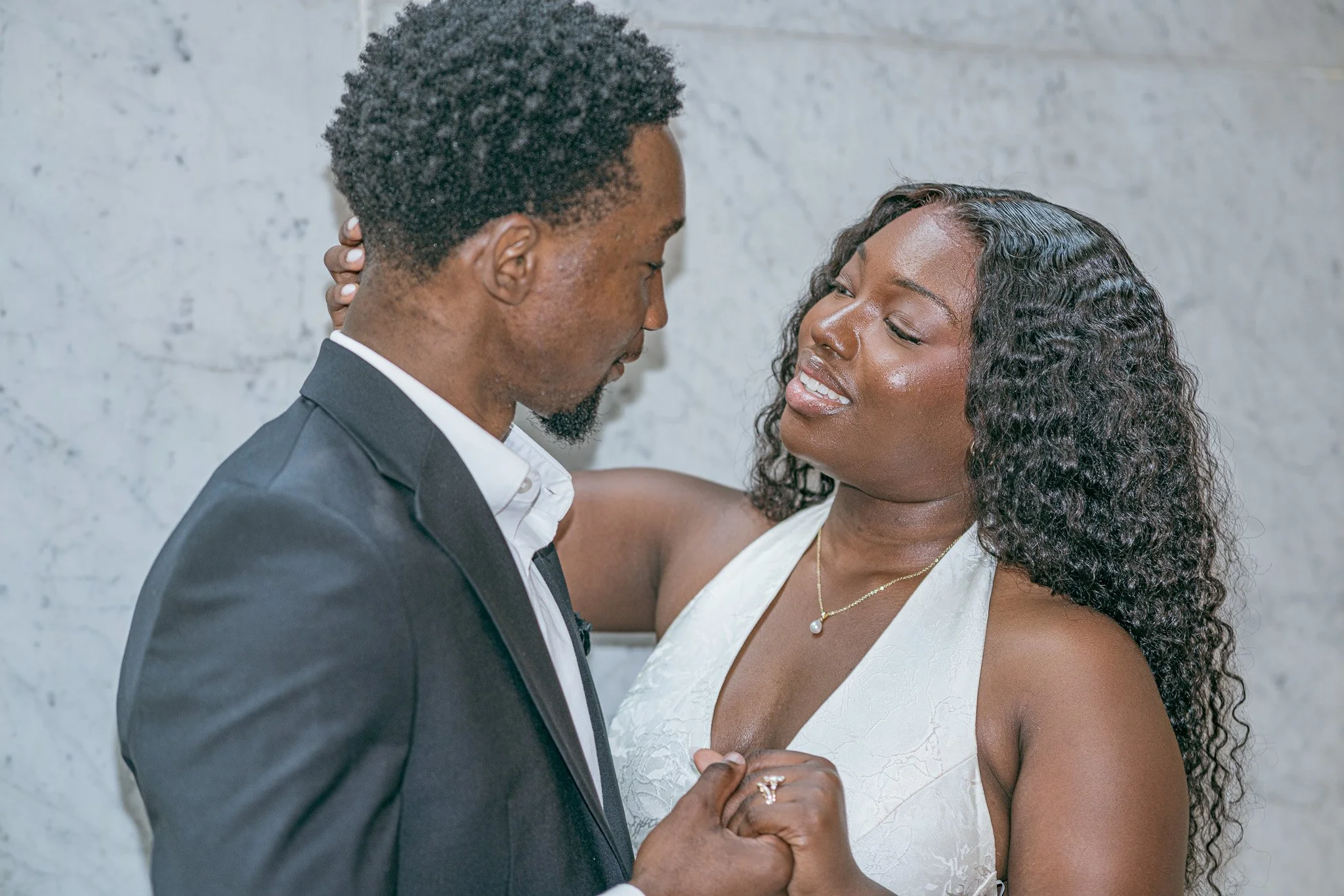 A couple sharing a dance, looking into each other's eyes, with the man wearing a dark suit and the woman in a white dress, against a gray textured wall.