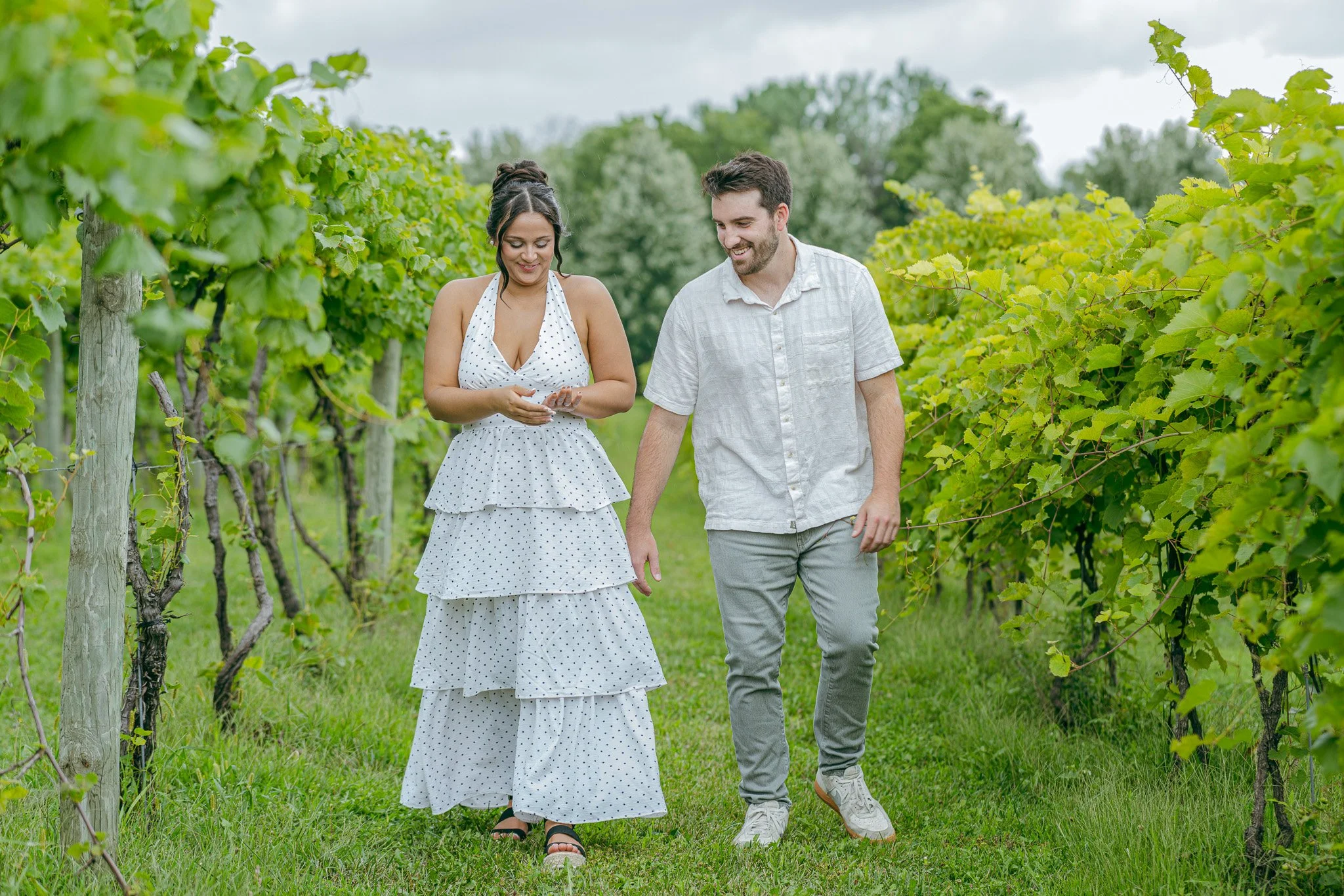 A woman in a white polka dot dress and man in a light gray shirt walking through a vineyard, smiling, surrounded by green grapevines.