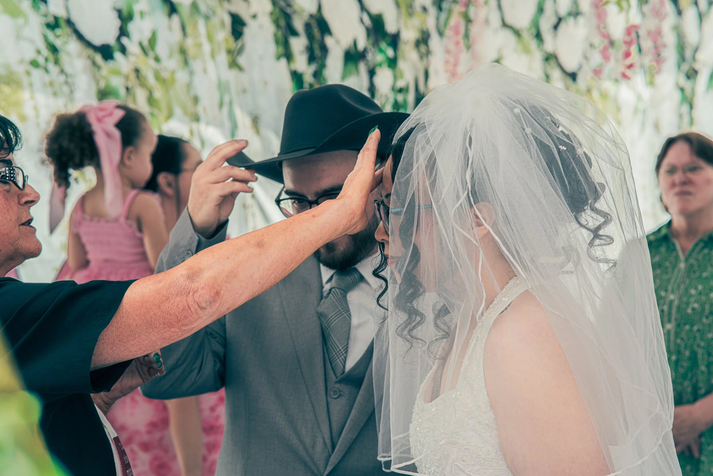 A wedding ceremony with a bride and groom under a canopy of greenery, with guests in attendance. The bride is wearing a wedding dress with a veil, and the groom is in a suit. Good moment captured as the groom adjusting his cowboy hat.