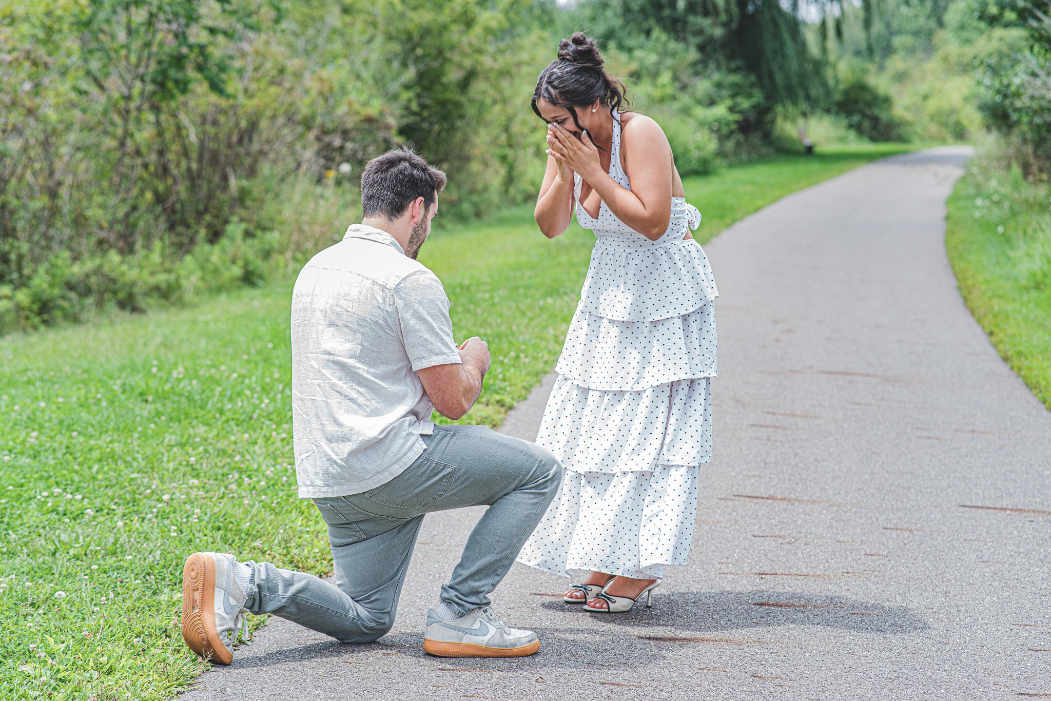 A man proposing marriage to a woman in a polka-dot dress on a park pathway, with the man kneeling and holding her hand, and the woman covering her mouth in surprise.