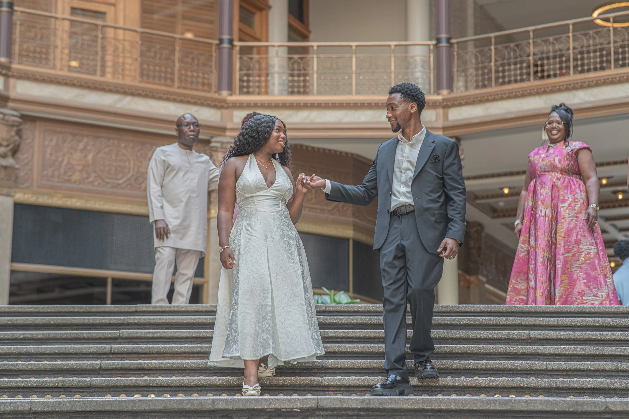 A wedding ceremony on the steps of a grand building with ornate woodwork, featuring a bride and groom holding hands and exchanging vows, with two women and a man standing in the background.