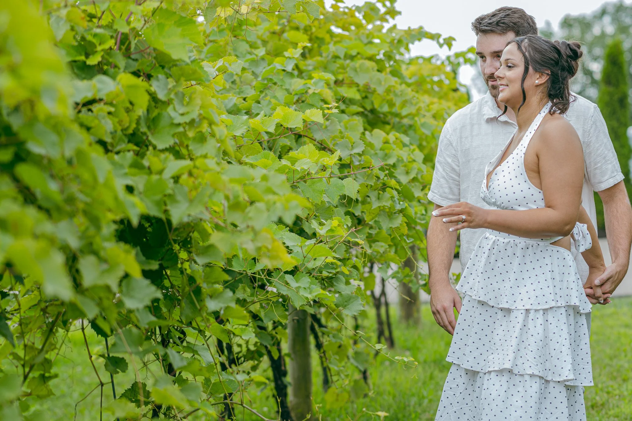 A couple holding hands and smiling while walking through a vineyard with green leaves.
