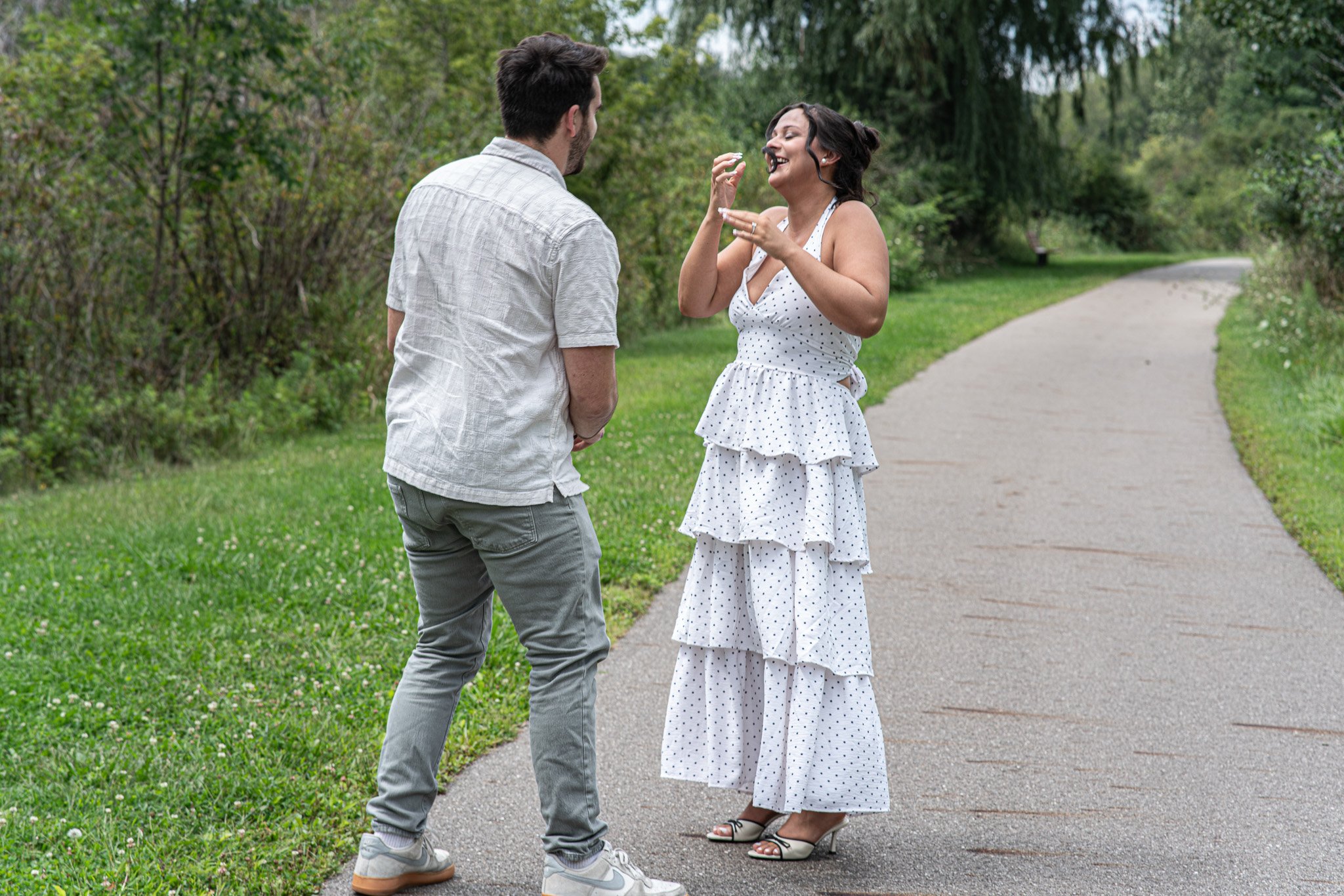 A man and woman stand on a park sidewalk, smiling and talking, with trees and greenery in the background.