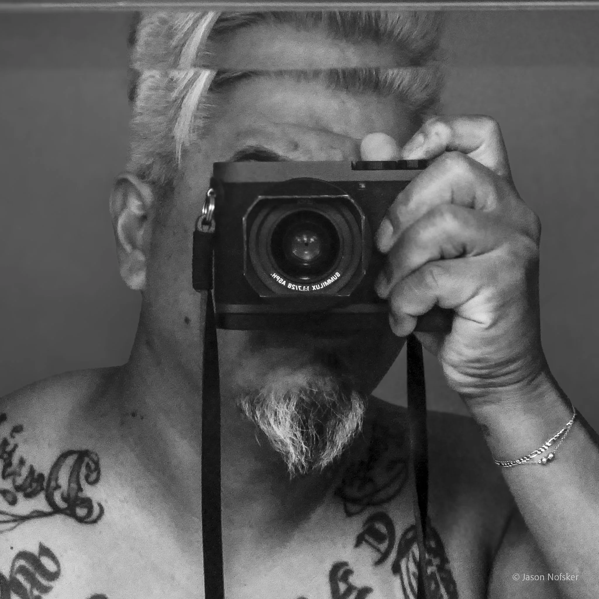 Close-up black and white photograph of a person taking a selfie with a camera, showing part of their face and hand holding the camera, with a focus on their facial hair and ear.