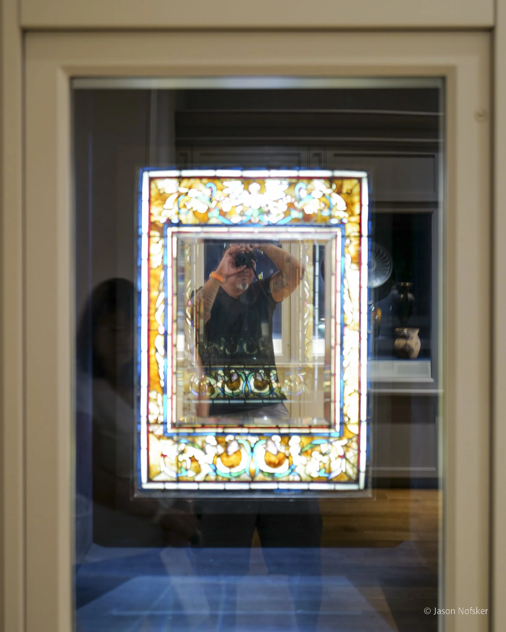 Reflection of a person taking a photo in a mirror with a stained glass window behind them.