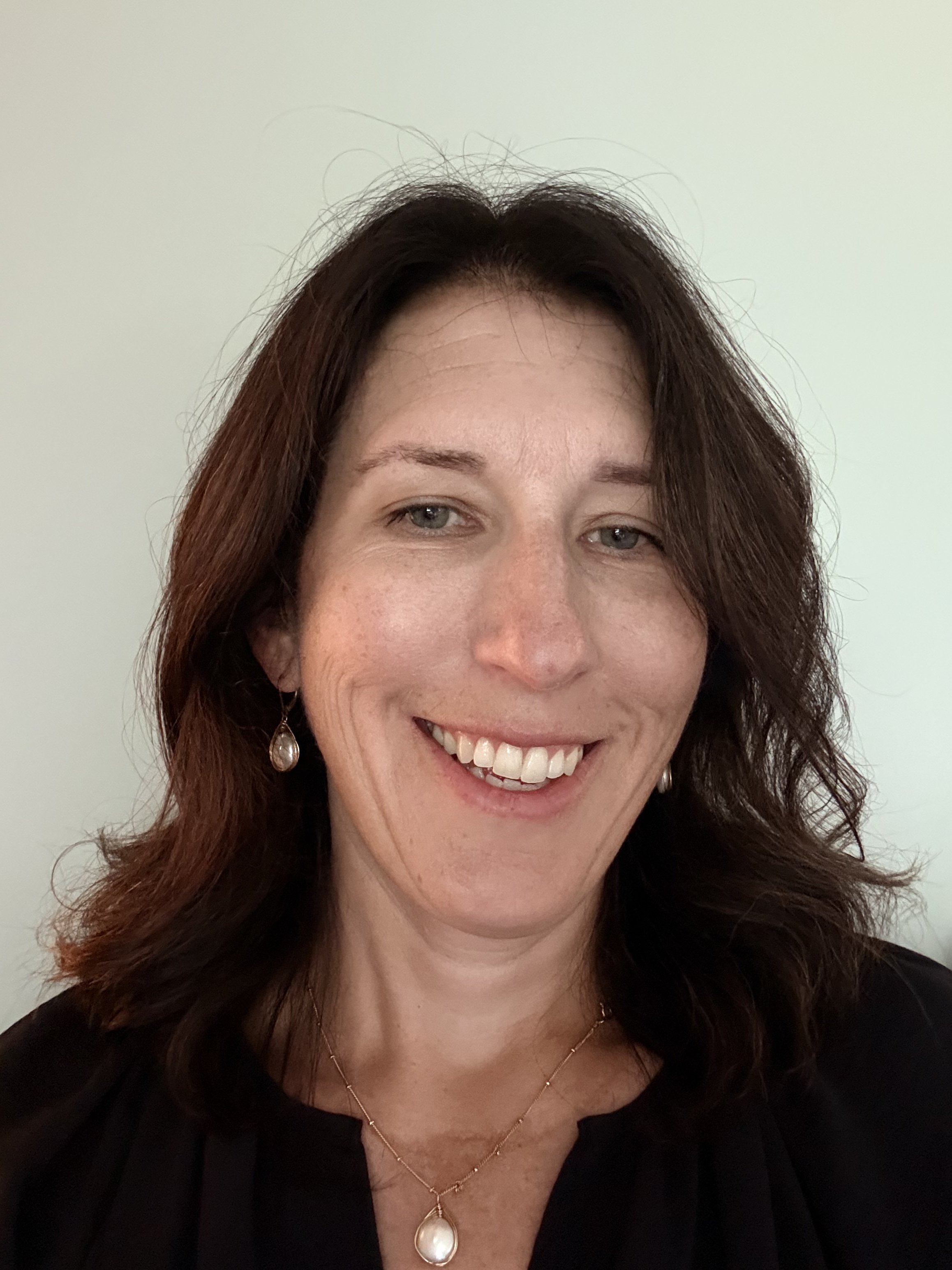 Close-up photo of a woman with dark brown wavy hair, smiling, wearing earrings and a pearl pendant necklace, against a plain light-colored background.
