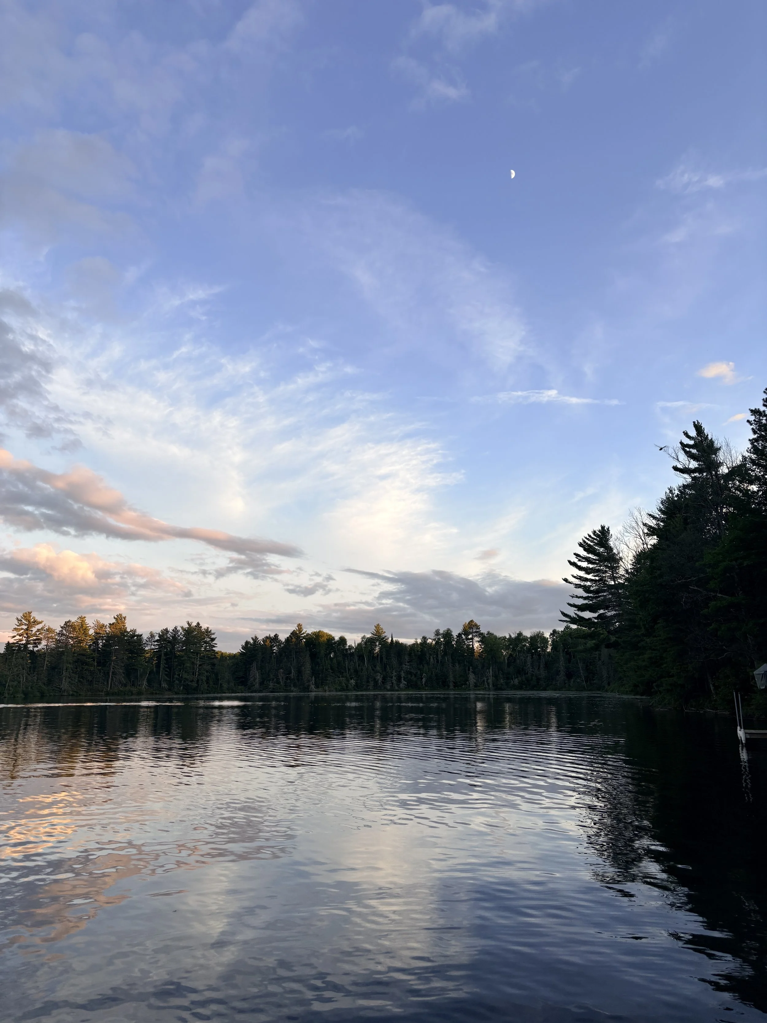 A serene landscape of a calm lake surrounded by trees under a blue sky with scattered clouds and a visible crescent moon.