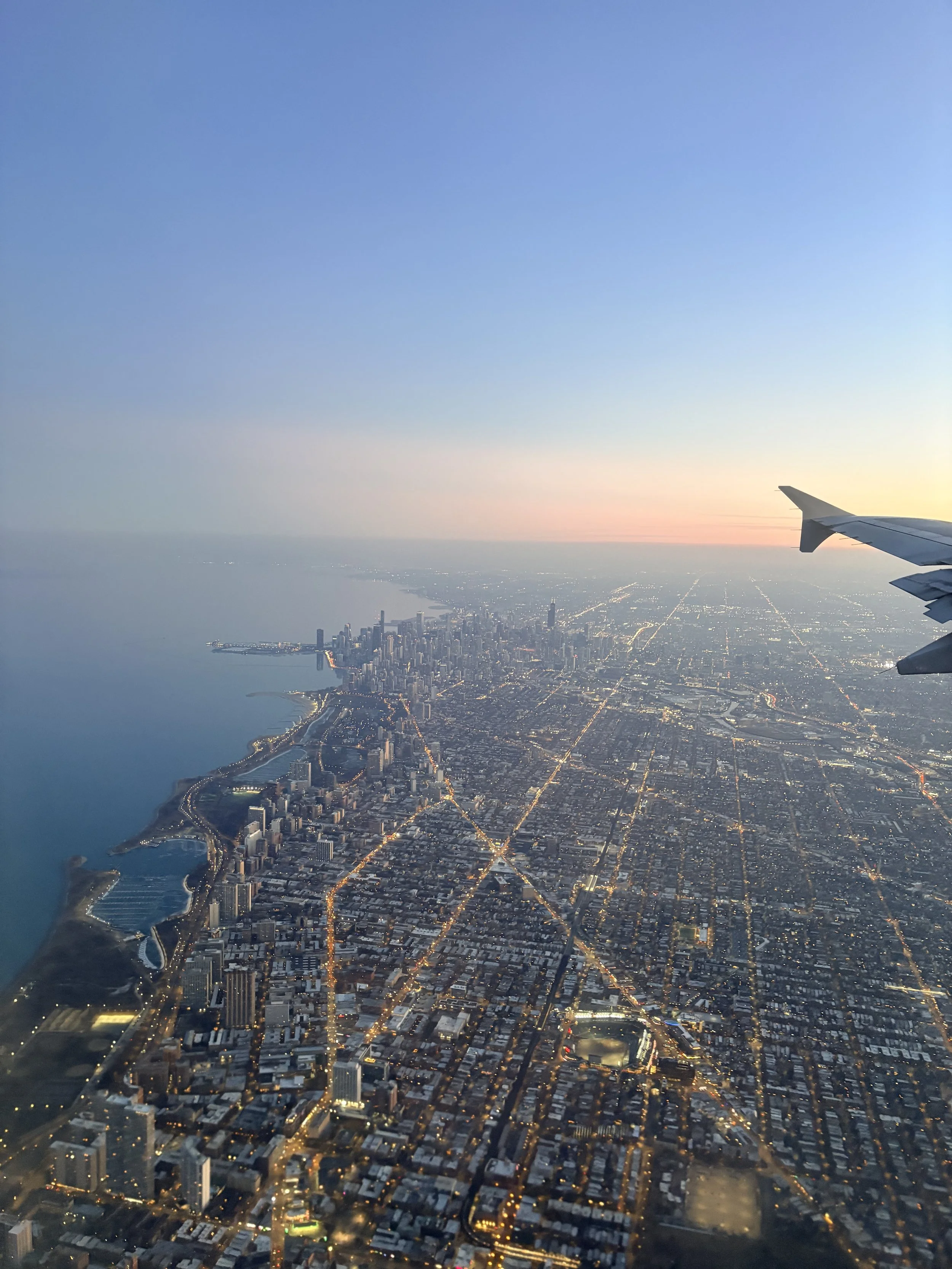 Aerial view of a cityscape at dusk with illuminated streets and tall buildings along the coastline, with city lights sparkling and the sky transitioning from sunset to night.