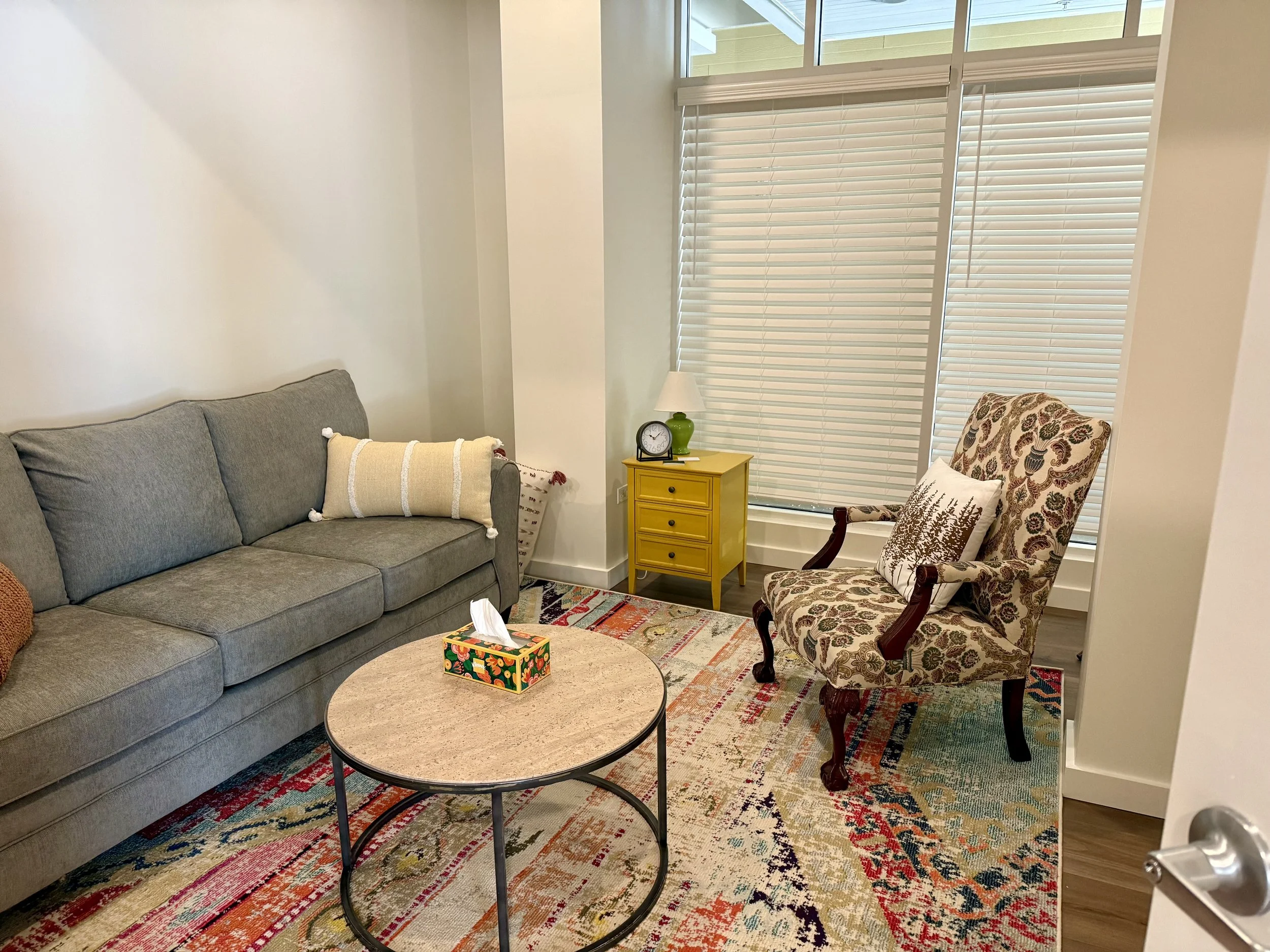 A cozy living room with a gray sofa adorned with colorful pillows, a round coffee table with a tissue box, a yellow side table with a green lamp and clock, a patterned armchair, a colorful area rug, and window blinds allowing natural light.