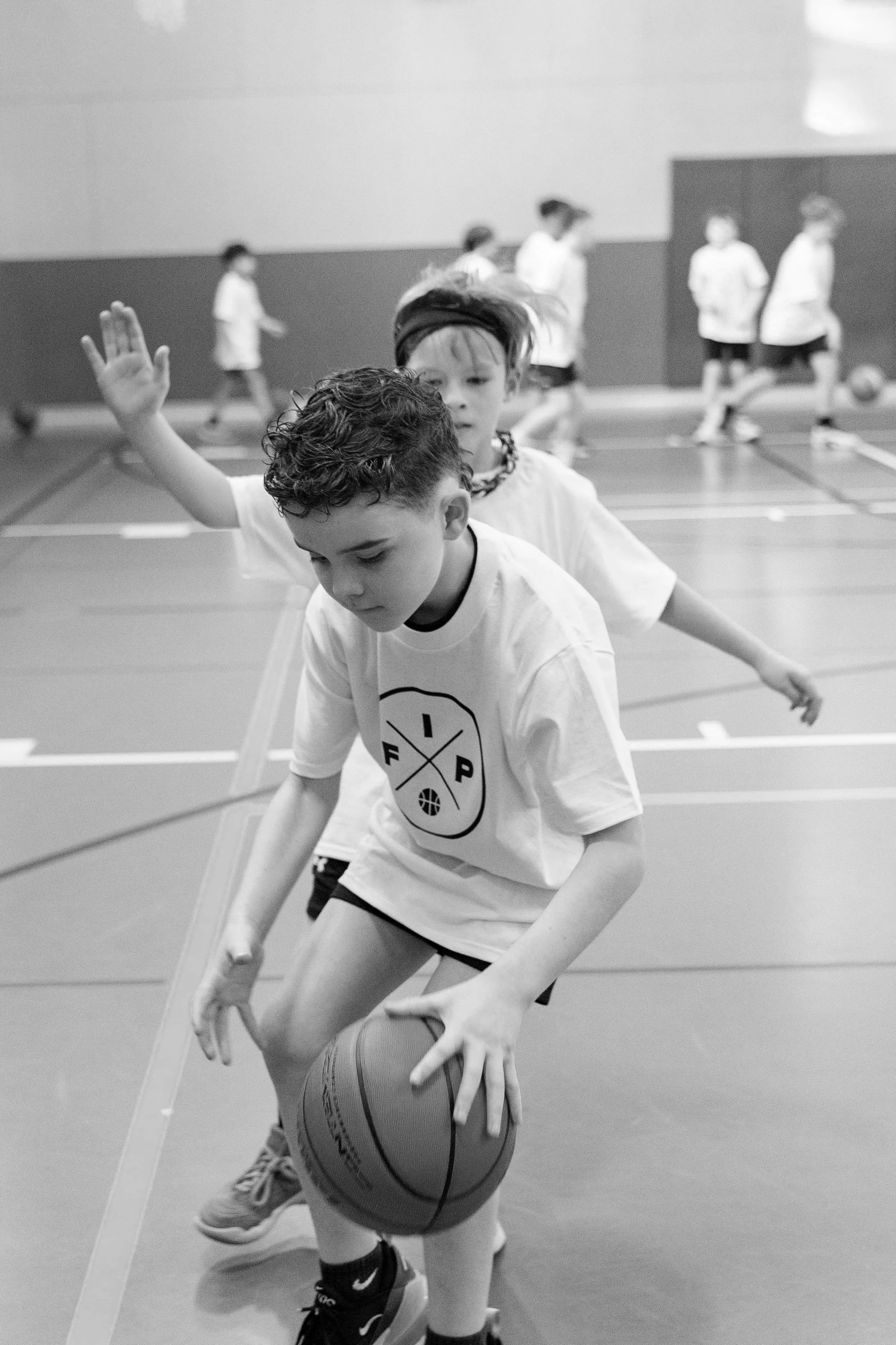 Children playing basketball in an indoor gym, one child in the foreground dribbling a basketball while others are in the background.