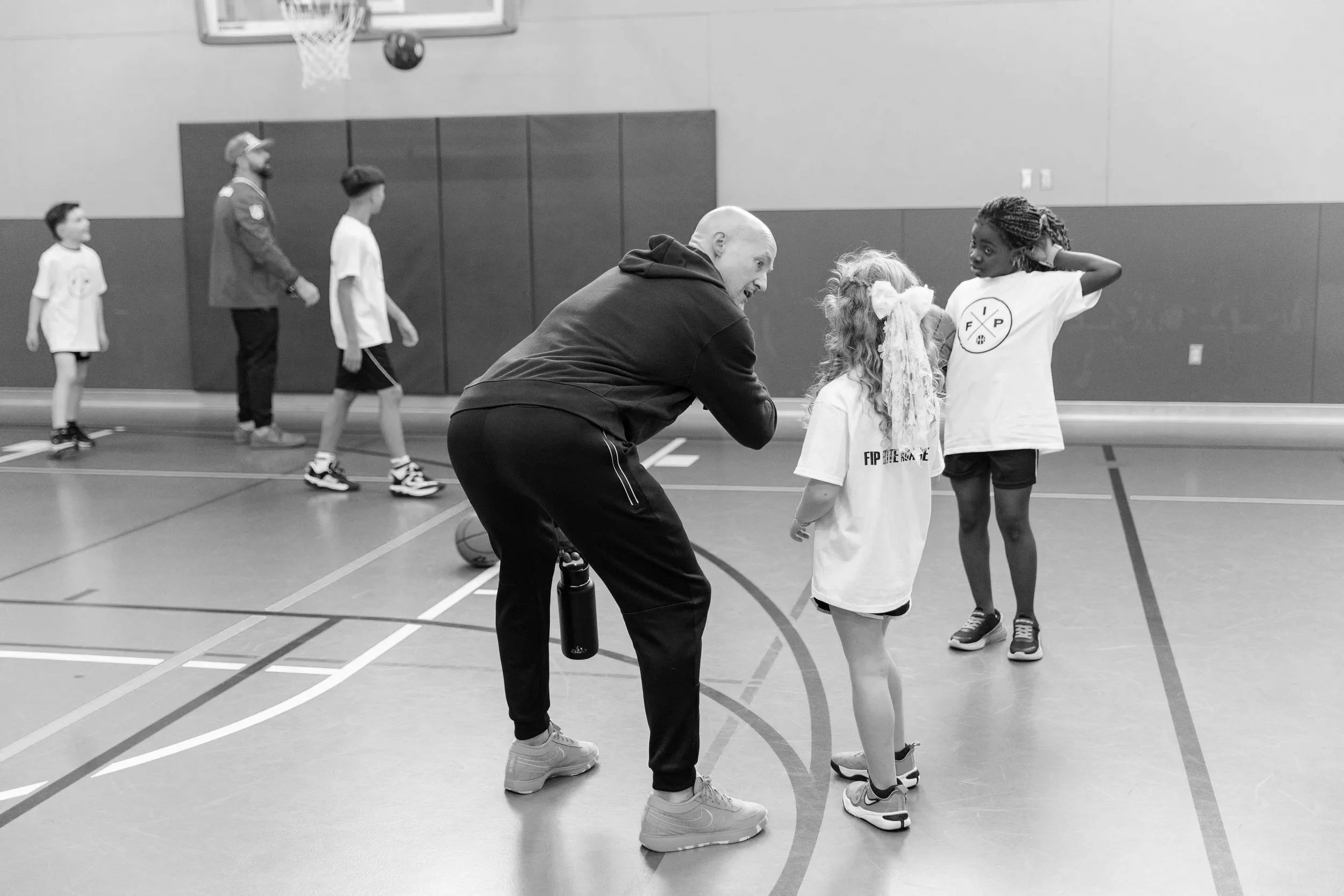 A coach talking to two young girls in a gym. The coach is crouching while the girls are standing and listening. In the background, three other children are walking across the gym, which has a basketball hoop and painted lines on the floor.