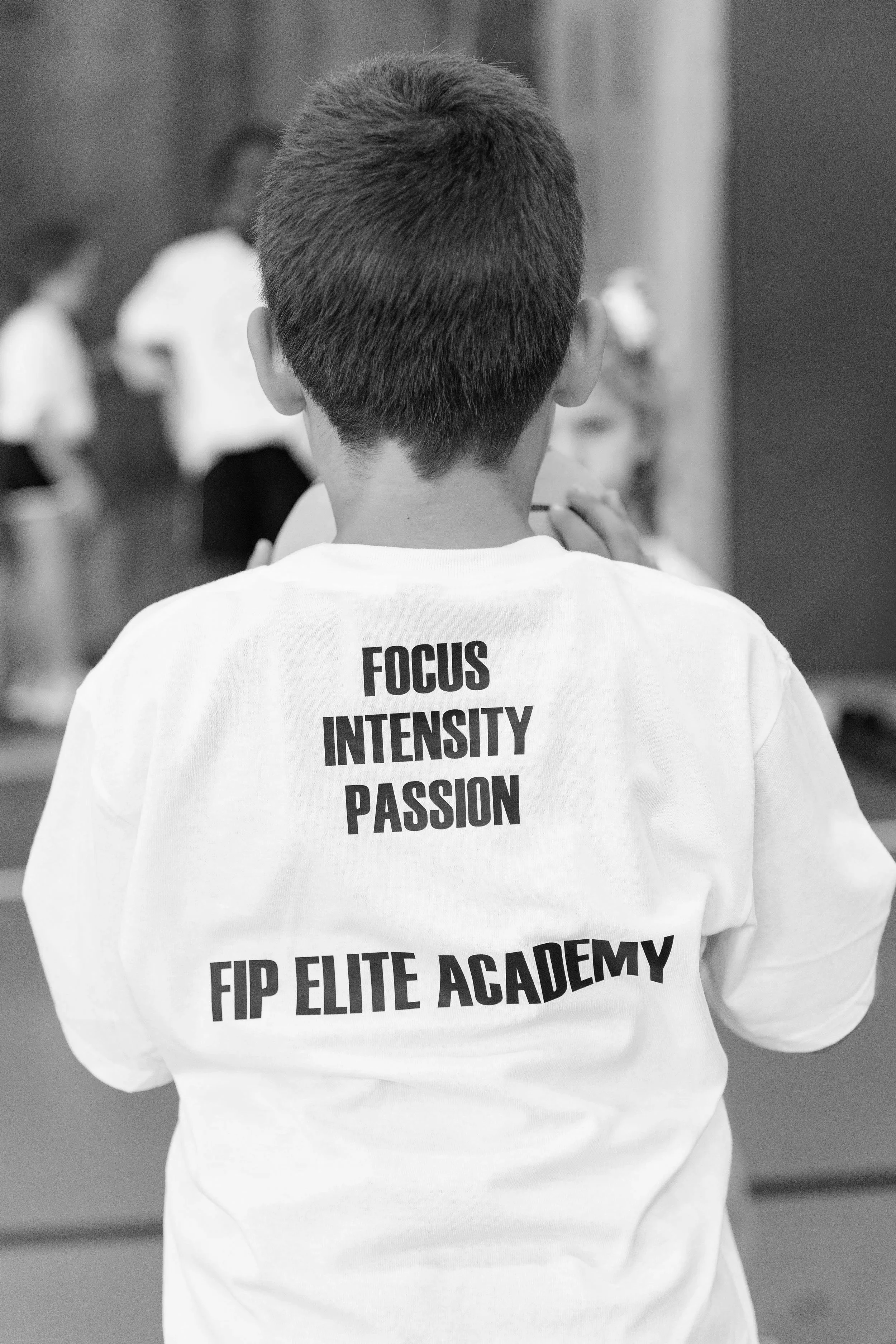 Back view of a person wearing a white t-shirt with the words "FOCUS, INTENSITY, PASSION, FIP ELITE ACADEMY" printed on it. The person has short hair and is in a gym or training setting, with other people blurred in the background.