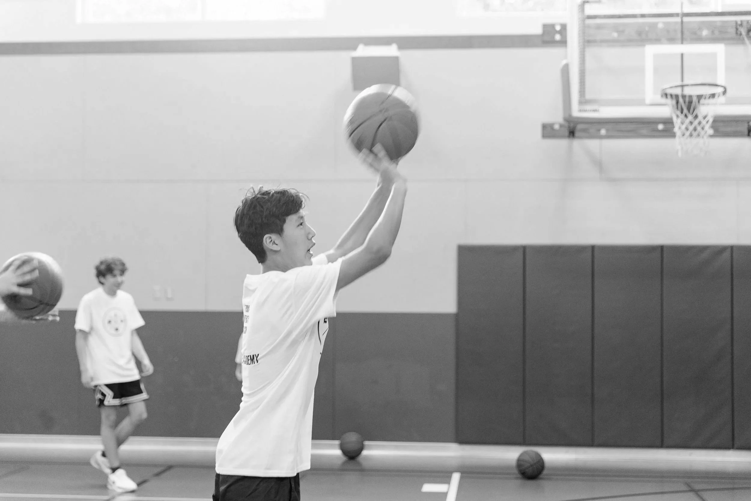 Young boy playing basketball in an indoor gym, preparing to shoot the ball towards the hoop, with other kids in the background.