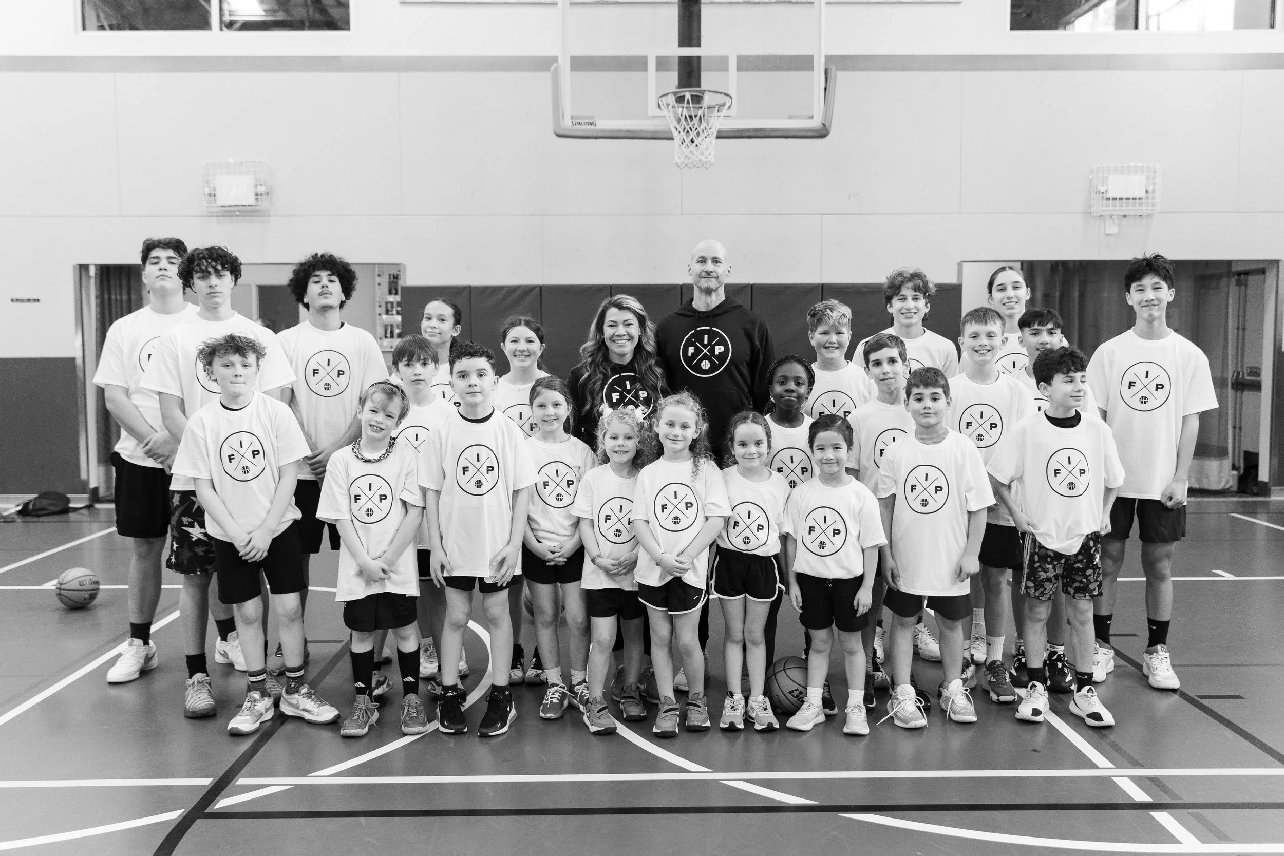 A group of children and two adult coaches standing on a basketball court in a gymnasium, all wearing matching t-shirts with a logo, with a basketball and a hoop visible in the background.