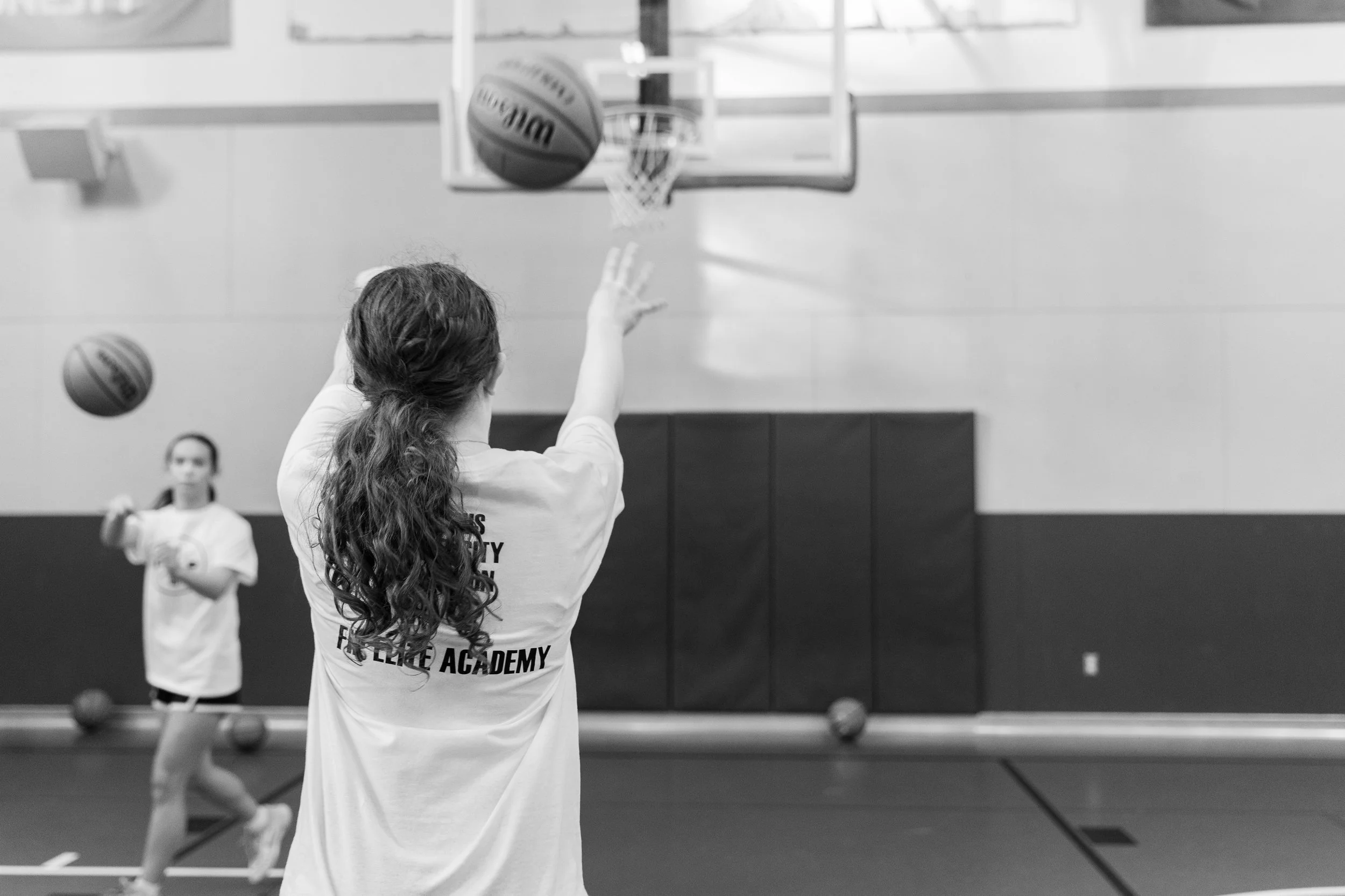 A girl with curly hair taking a basketball shot in an indoor gym. Two other girls are in the background, with a basketball court, hoops, and gymnasium walls visible.