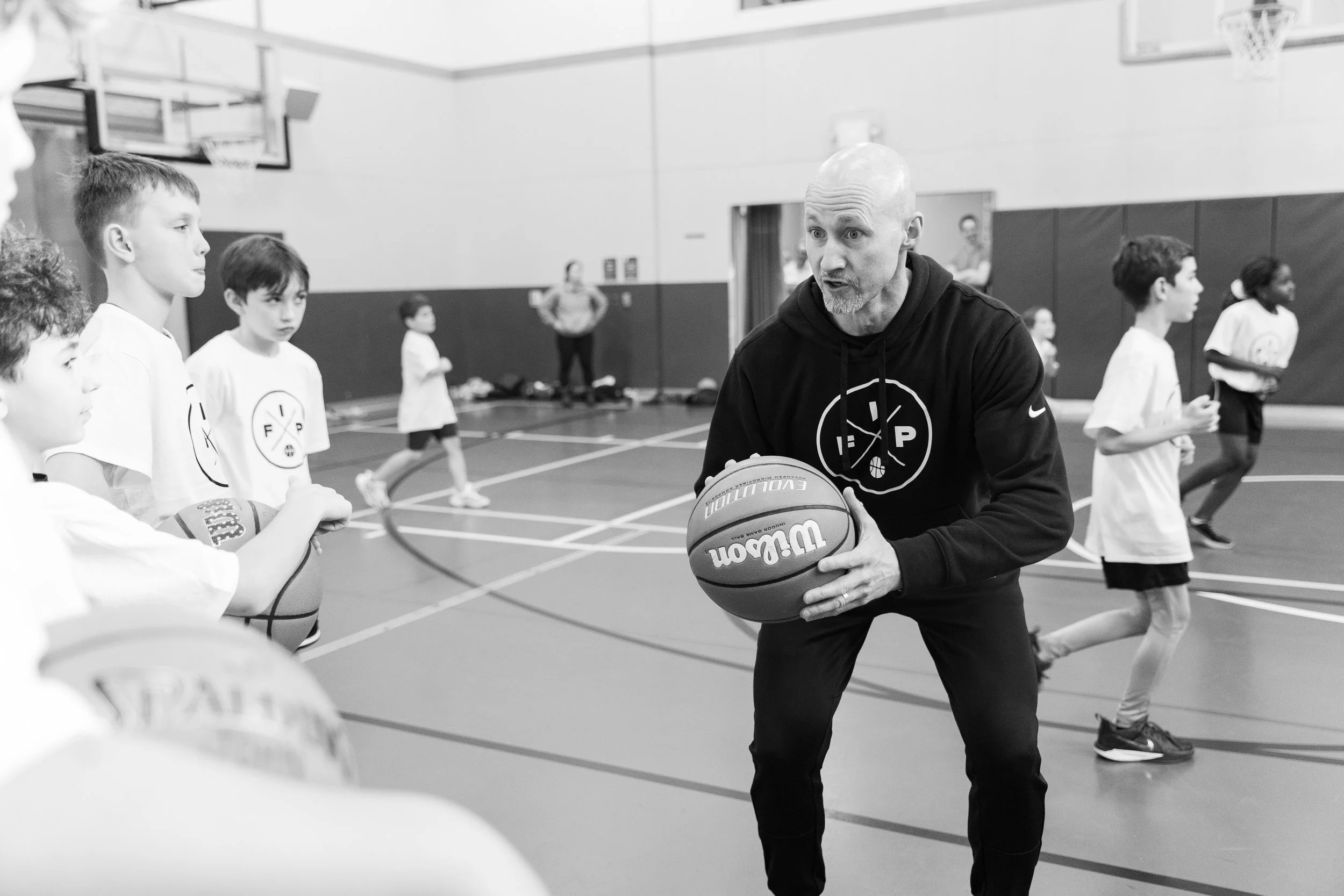 A coach instructs young children in basketball drills inside a gymnasium, with some children holding basketballs and others running.
