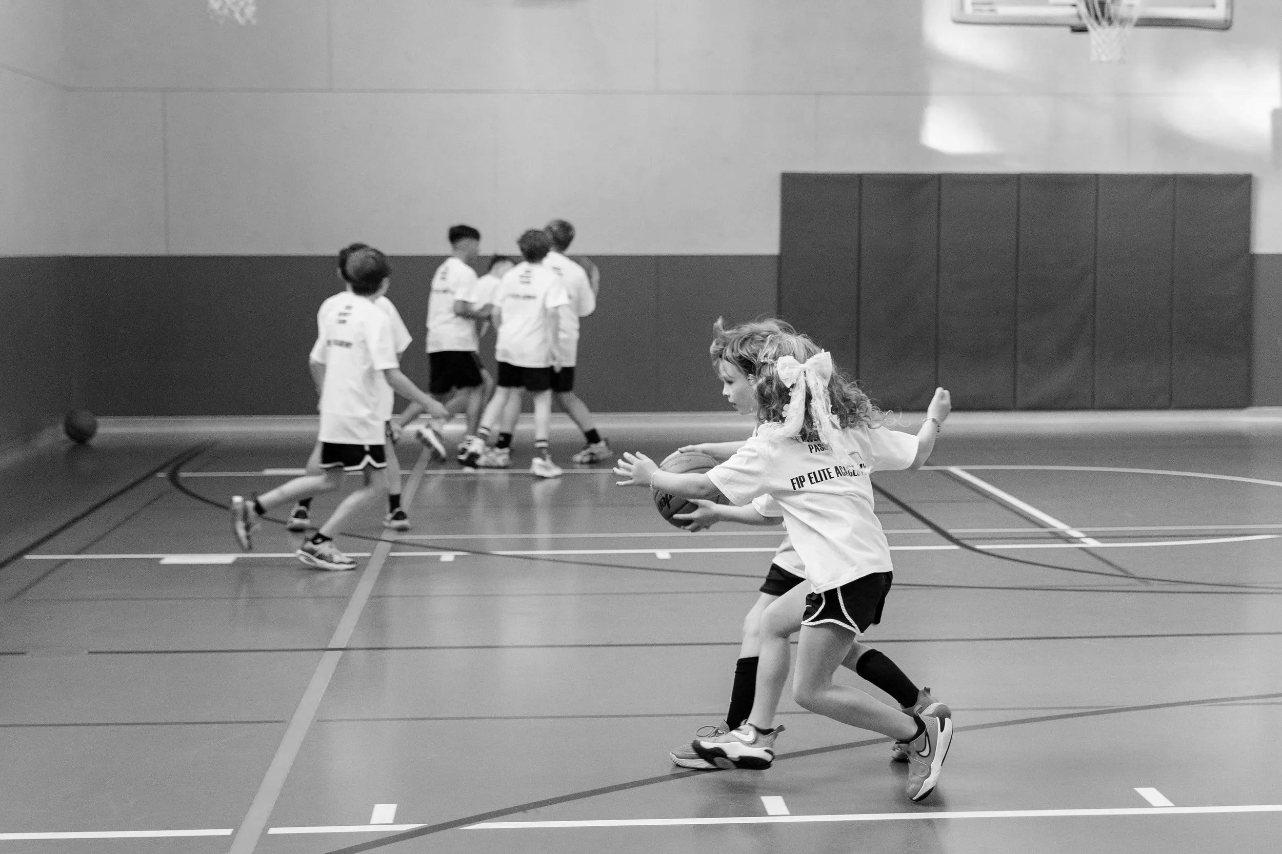 Children playing basketball indoors, with some running and one girl preparing to pass the ball, wearing sports jerseys and shorts.