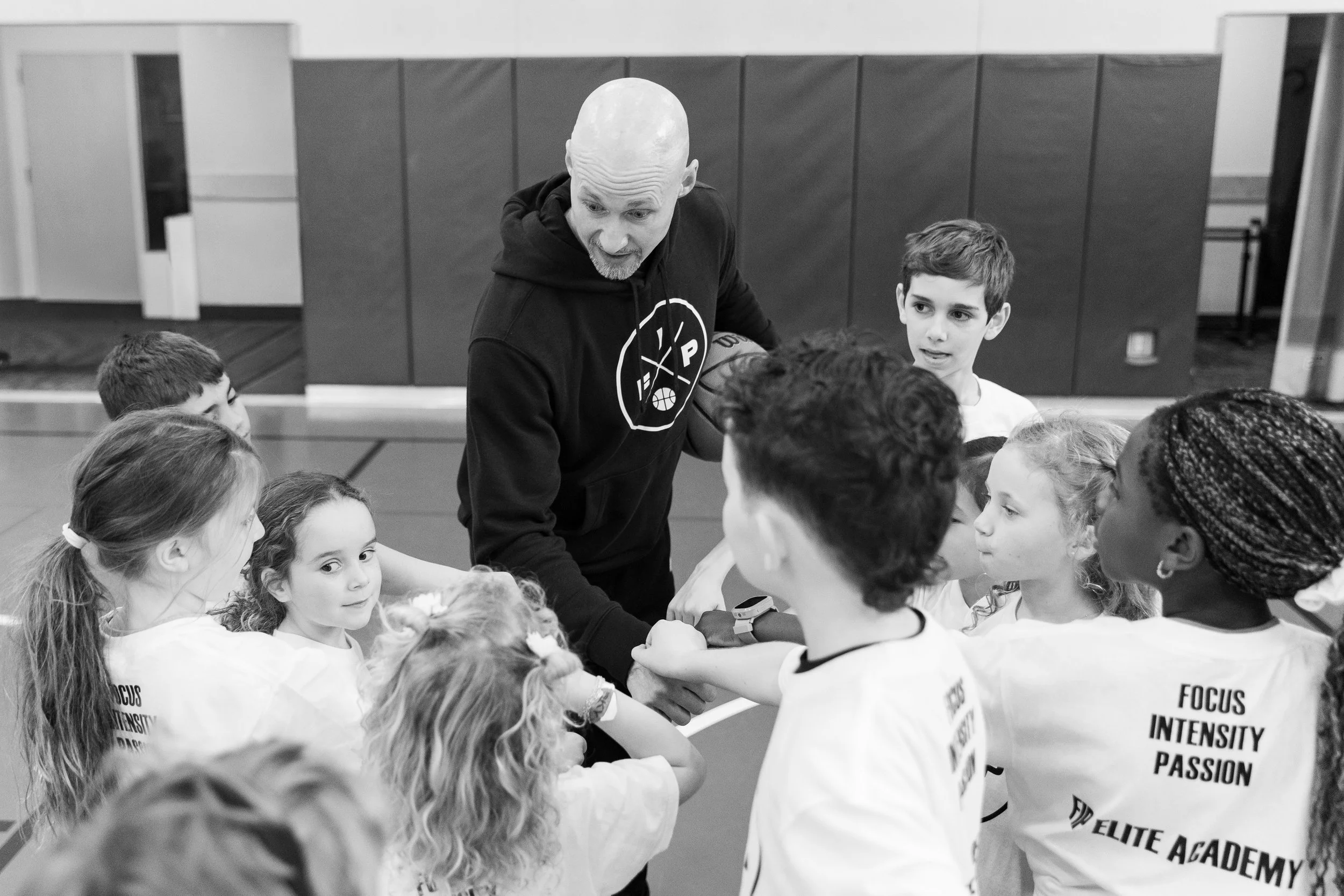 An adult male coach holding a basketball and talking to a group of children in a gymnasium, some of whom are wearing T-shirts with words like "Focus," "Intensity," and "Passion" on the back.