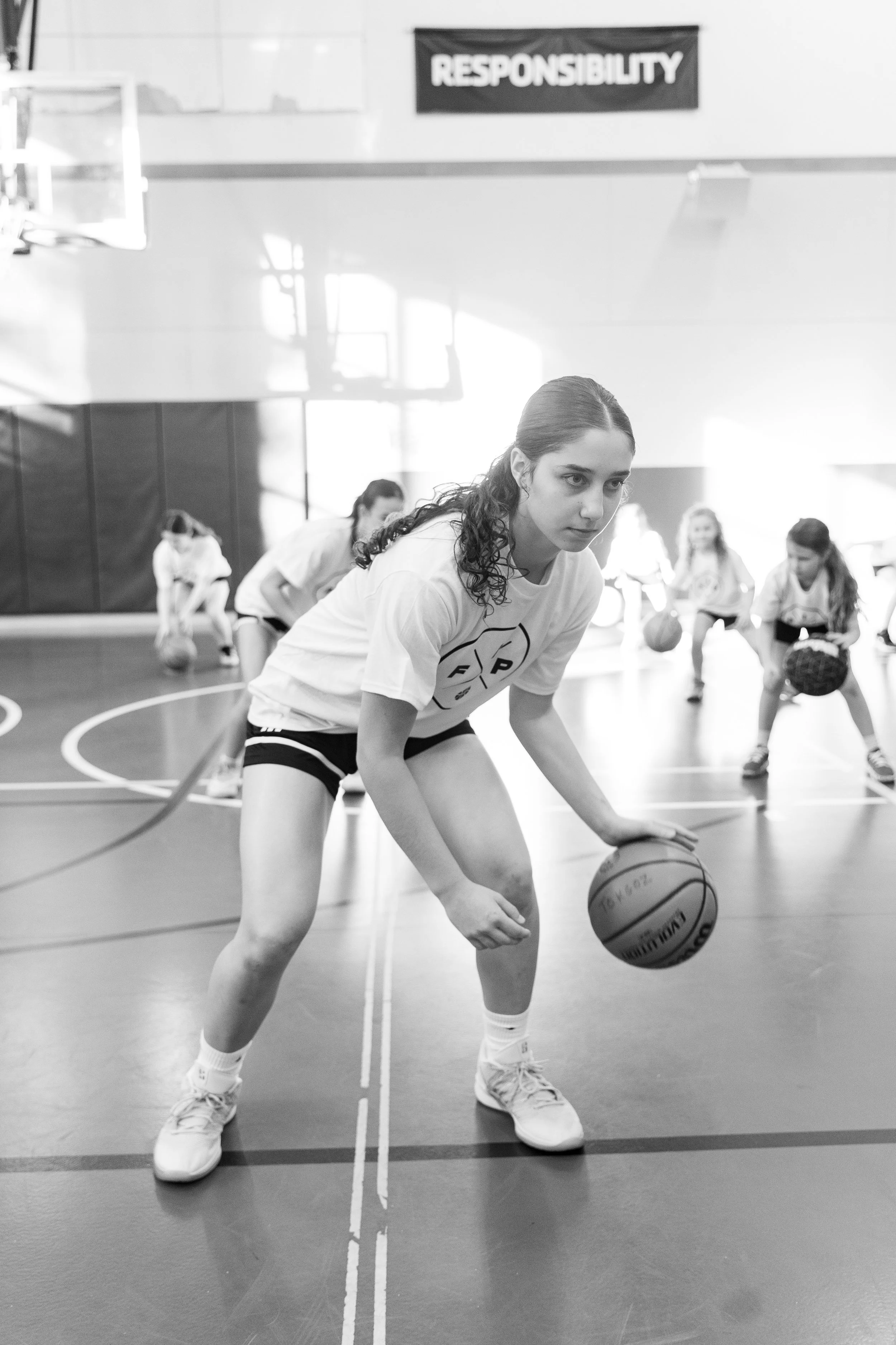 A young woman with curly hair dribbling a basketball on a gymnasium court with other children practicing in the background. There is a sign reading 'RESPONSIBILITY' on the wall.