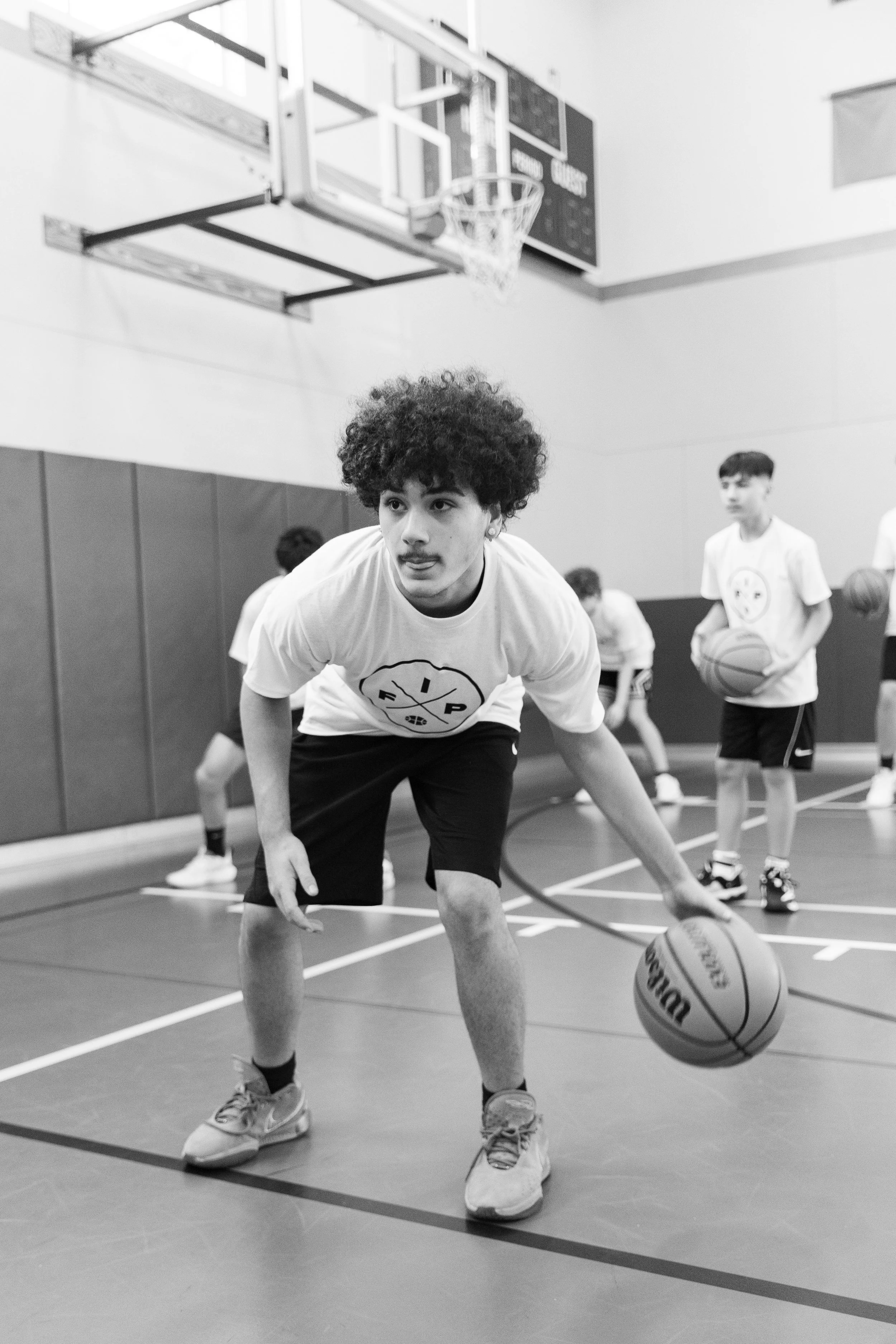 Young man in a gymnasium, playing basketball, bending down with a basketball near his feet, other kids in the background holding basketballs.