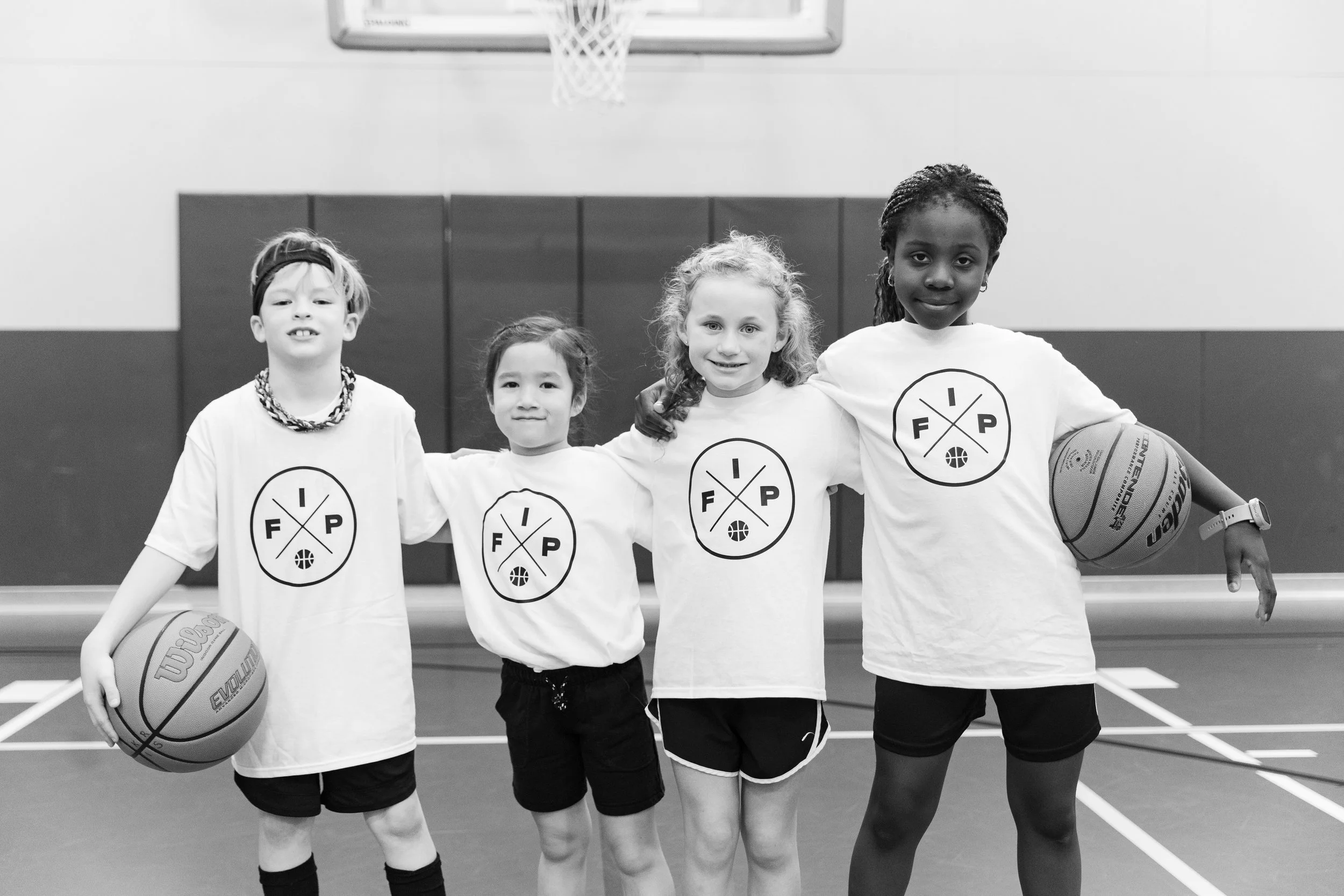 Four young girls standing in a gymnasium, dressed in basketball uniforms with a logo on their shirts, smiling, with two holding basketballs. There is a basketball hoop and scoreboard in the background.