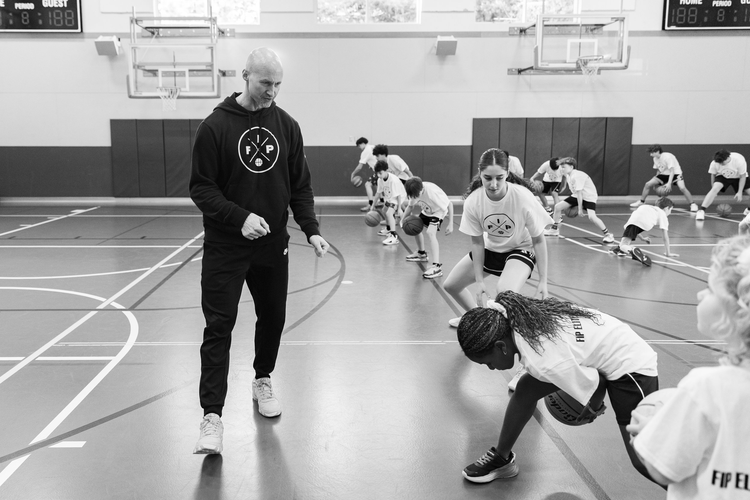 A basketball coach instructs young players during a practice session in a gymnasium, with multiple children dribbling basketballs in the background.