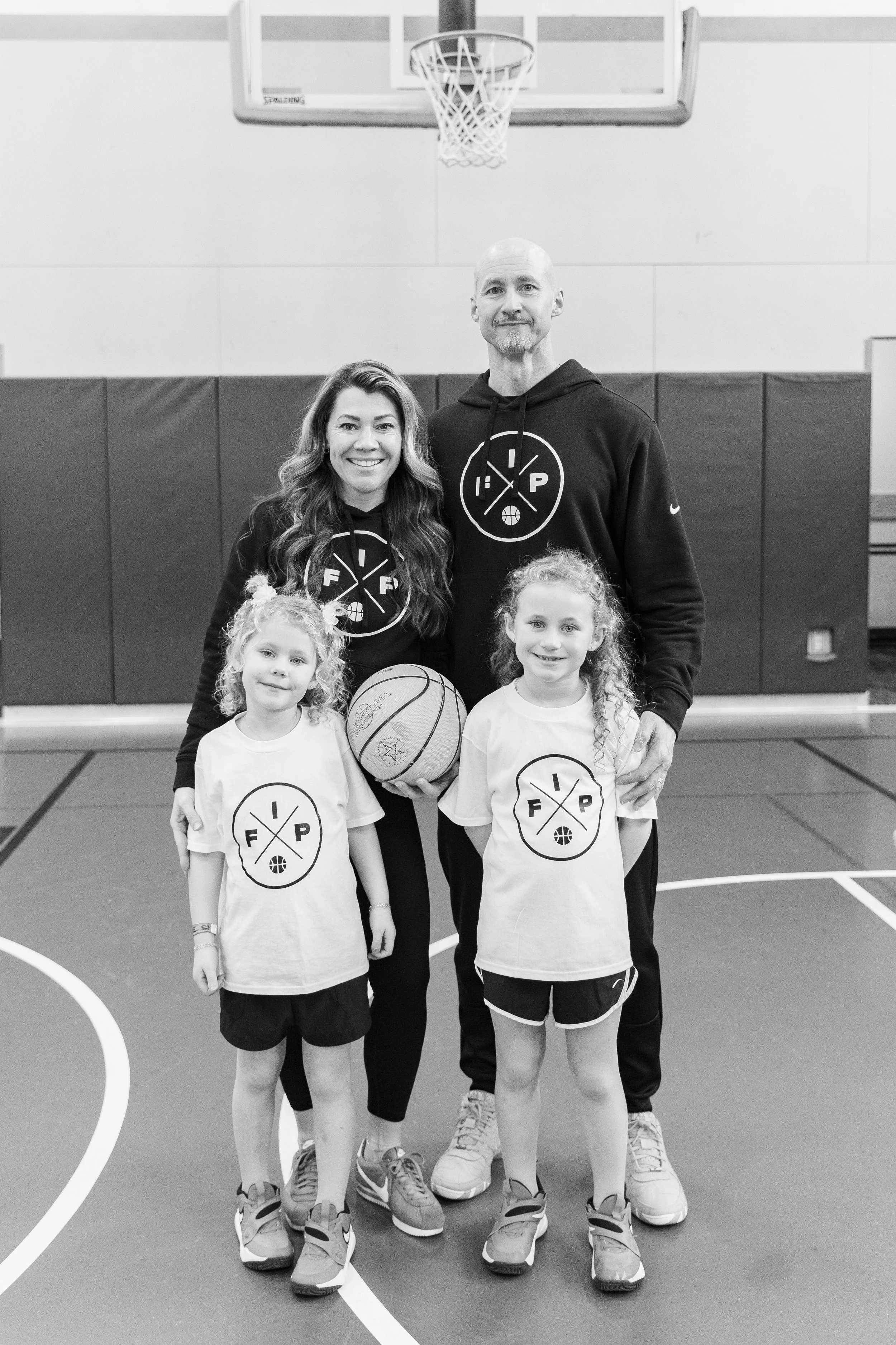 A group of four people, two children and two adults, standing on a basketball court inside a gymnasium, all smiling at the camera. The children are wearing white T-shirts with a logo featuring a crossed basketball and text that reads 'FIP' and 'EXP.' The adults are dressed in hoodies with the same logo.