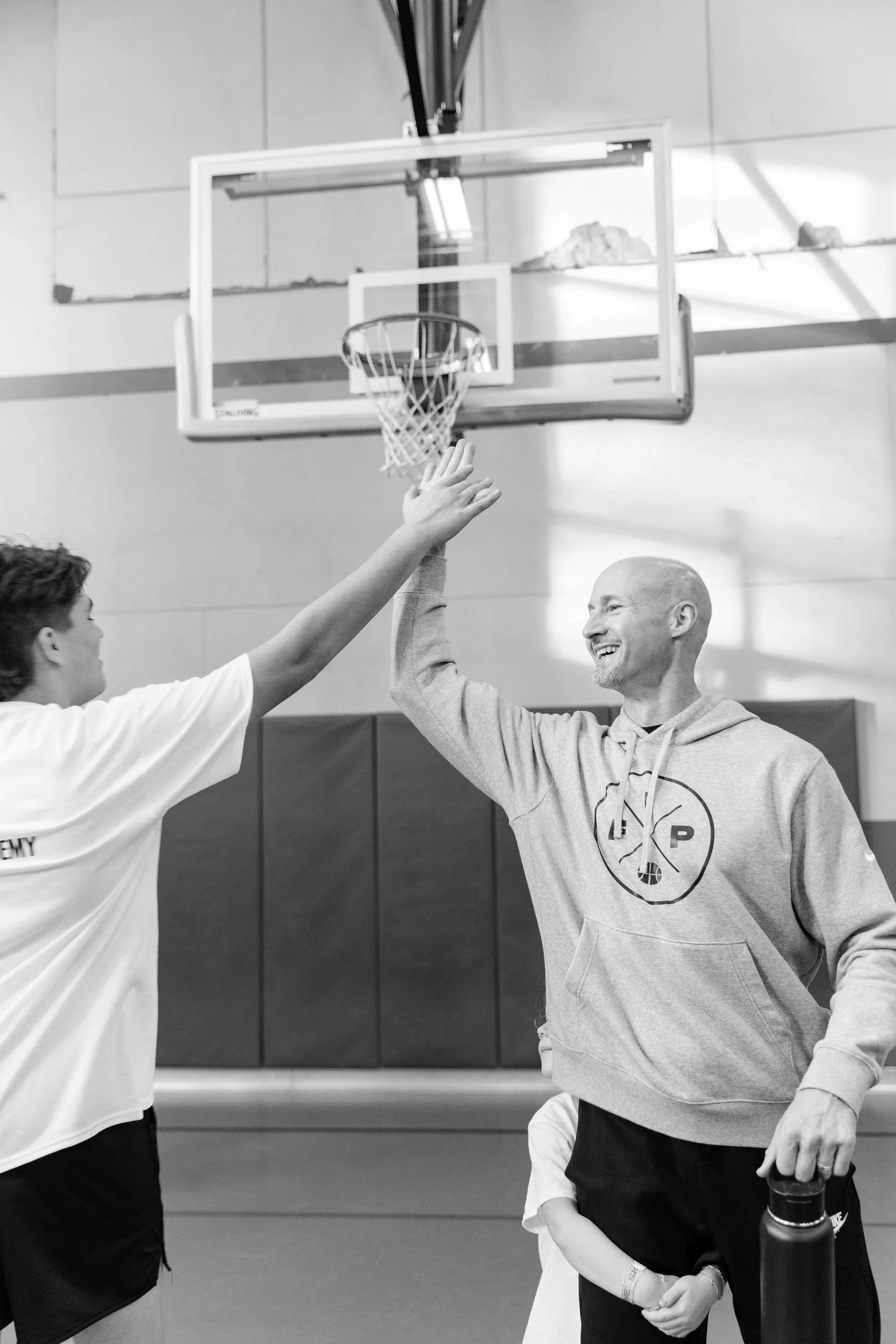 A basketball hoop in an indoor gym with two people giving each other a high five. One person is holding a water bottle and wearing a hoodie, and the other person is wearing a T-shirt.