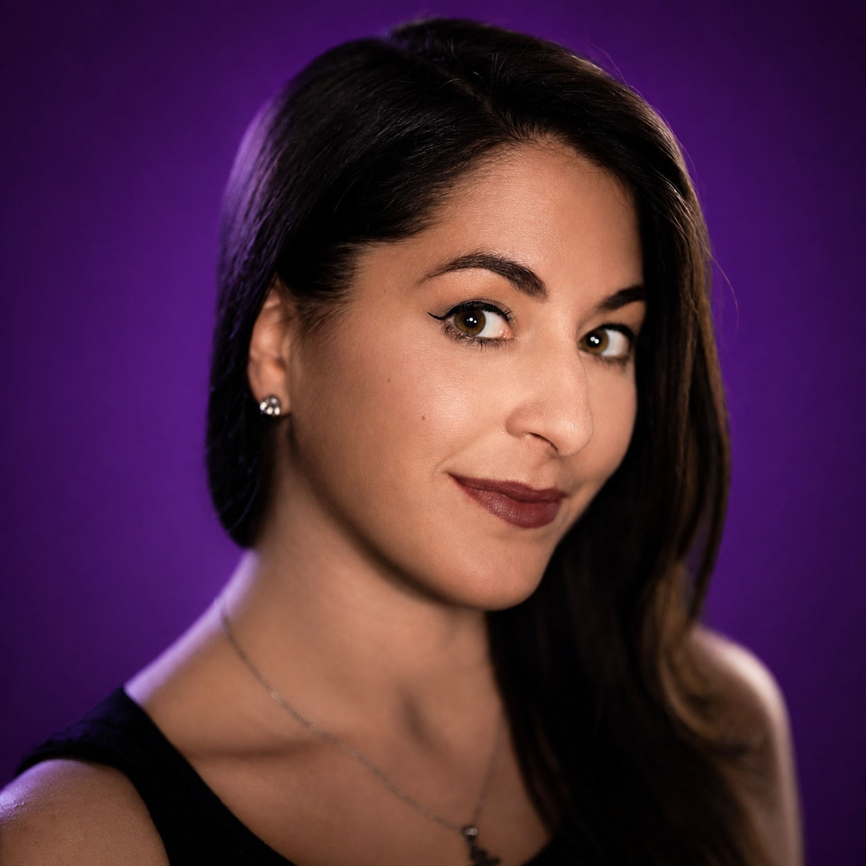 Close-up portrait of a woman with dark brown hair, brown eyes, and a subtle smile, wearing earrings and a black top against a purple background.