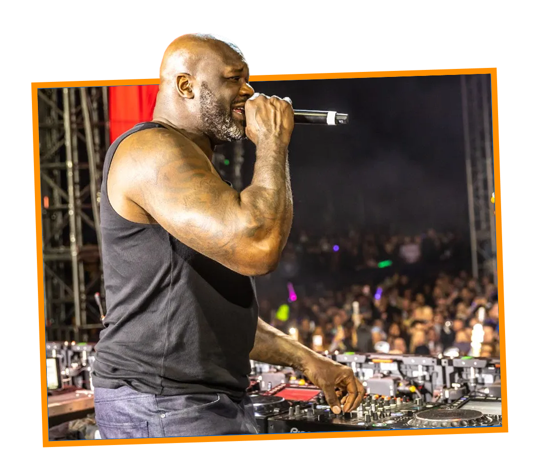 A DJ performing at a concert, holding a microphone and standing behind a mixing console with a crowd in the background.