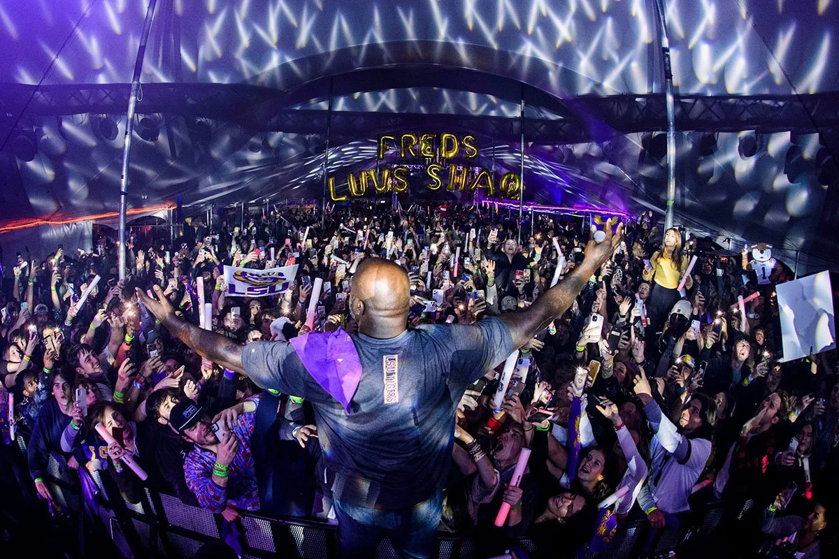 A performer on stage with arms raised facing a crowd at a concert or festival under a lit tent with decorative lighting. The crowd is waving glow sticks and taking photos. Signs and banners are visible in the audience.