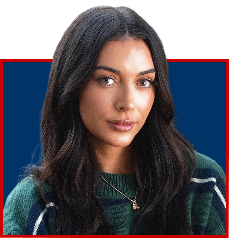 A young woman with long dark hair and fair skin looks at the camera, wearing a green and navy sweater with white stripes and a gold necklace, standing against a blue background.