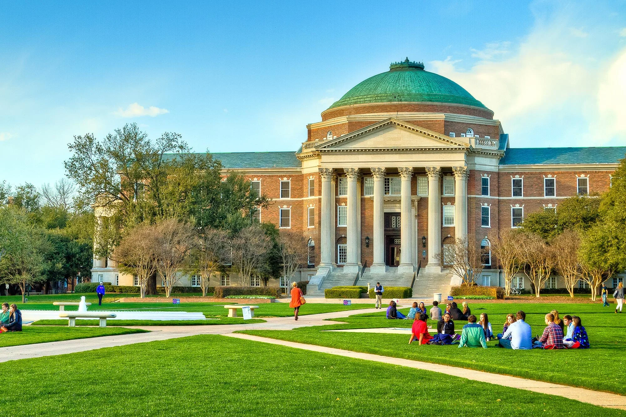 People sitting and walking on green lawn in front of a large brick building with a green dome and rows of columns, surrounded by trees under a blue sky.