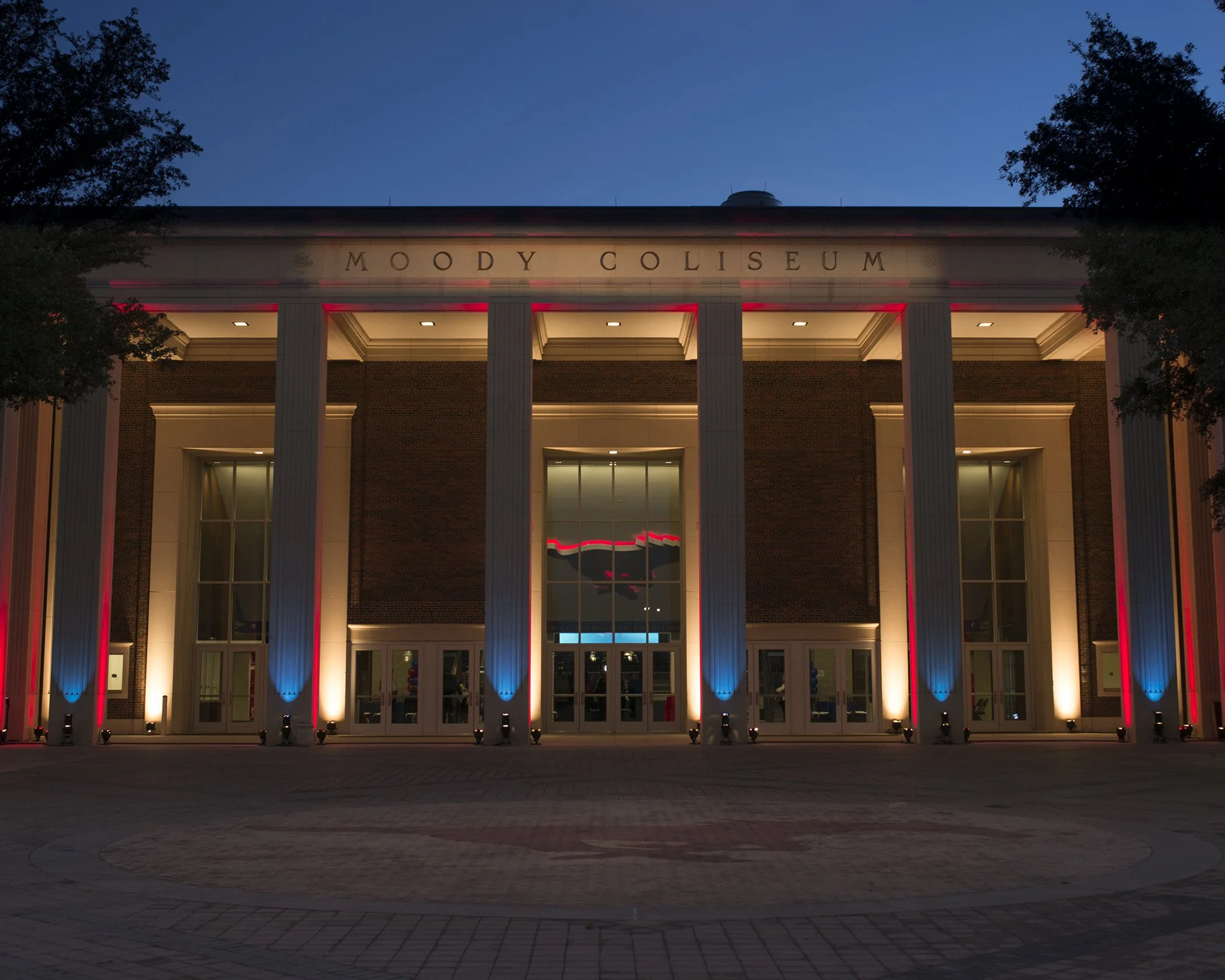 The Moody Coliseum is illuminated in red, white, and blue lights at night, with its name displayed above the entrance, framed by trees.