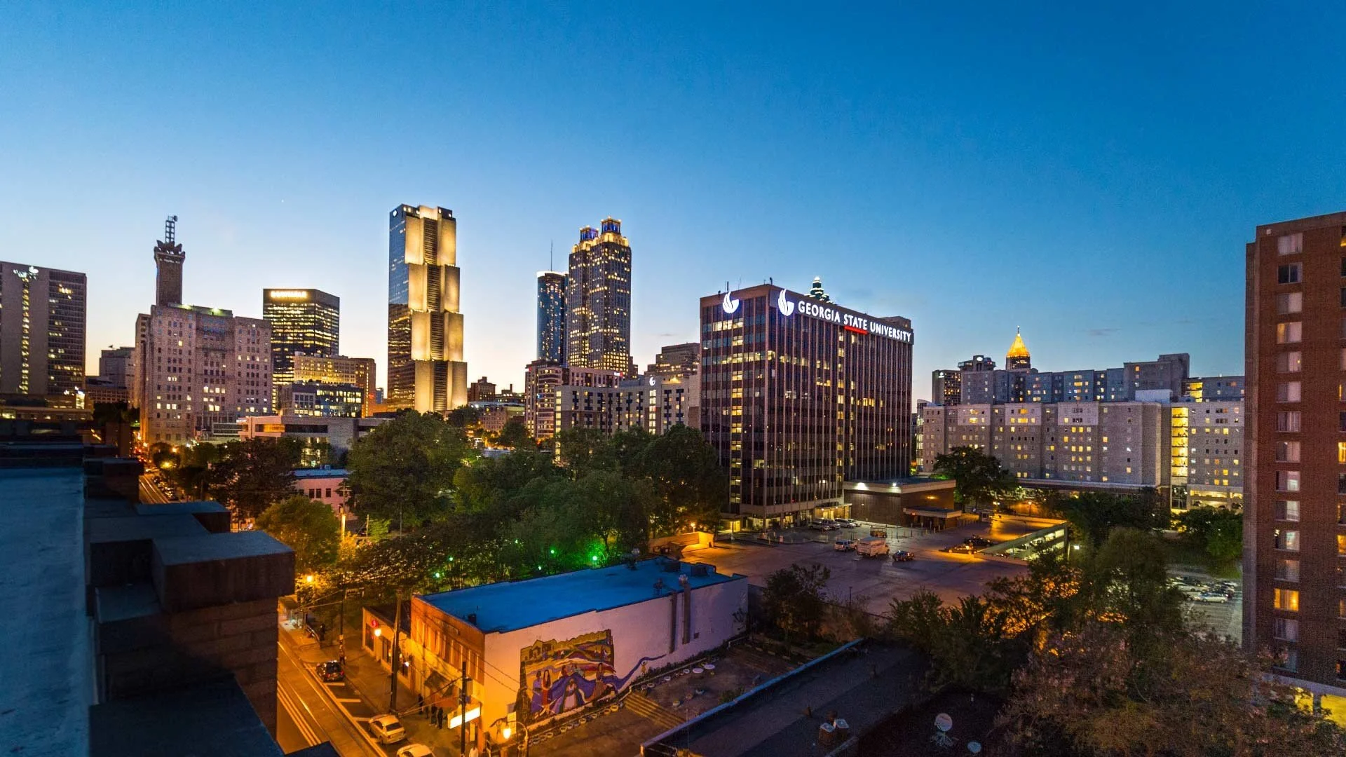 City skyline at dusk with tall buildings including Georgia State University sign, illuminated windows, trees, streets, and parked cars.