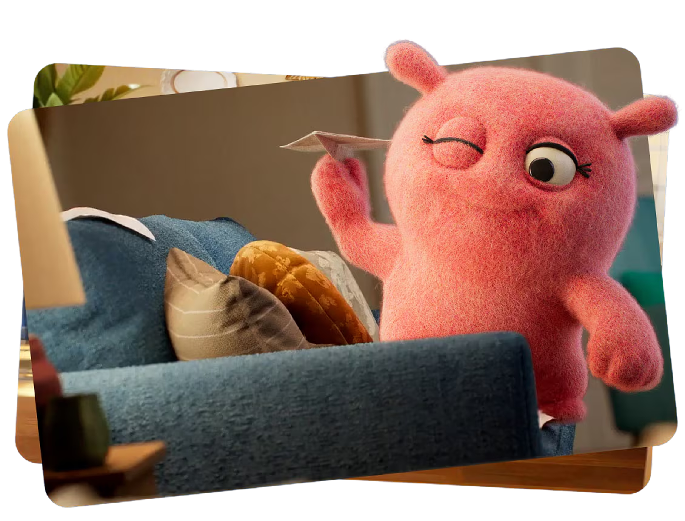 A pink, plush toy character with one eye open and the other eye closed, holding a note or paper, seen in a cozy indoor setting.
