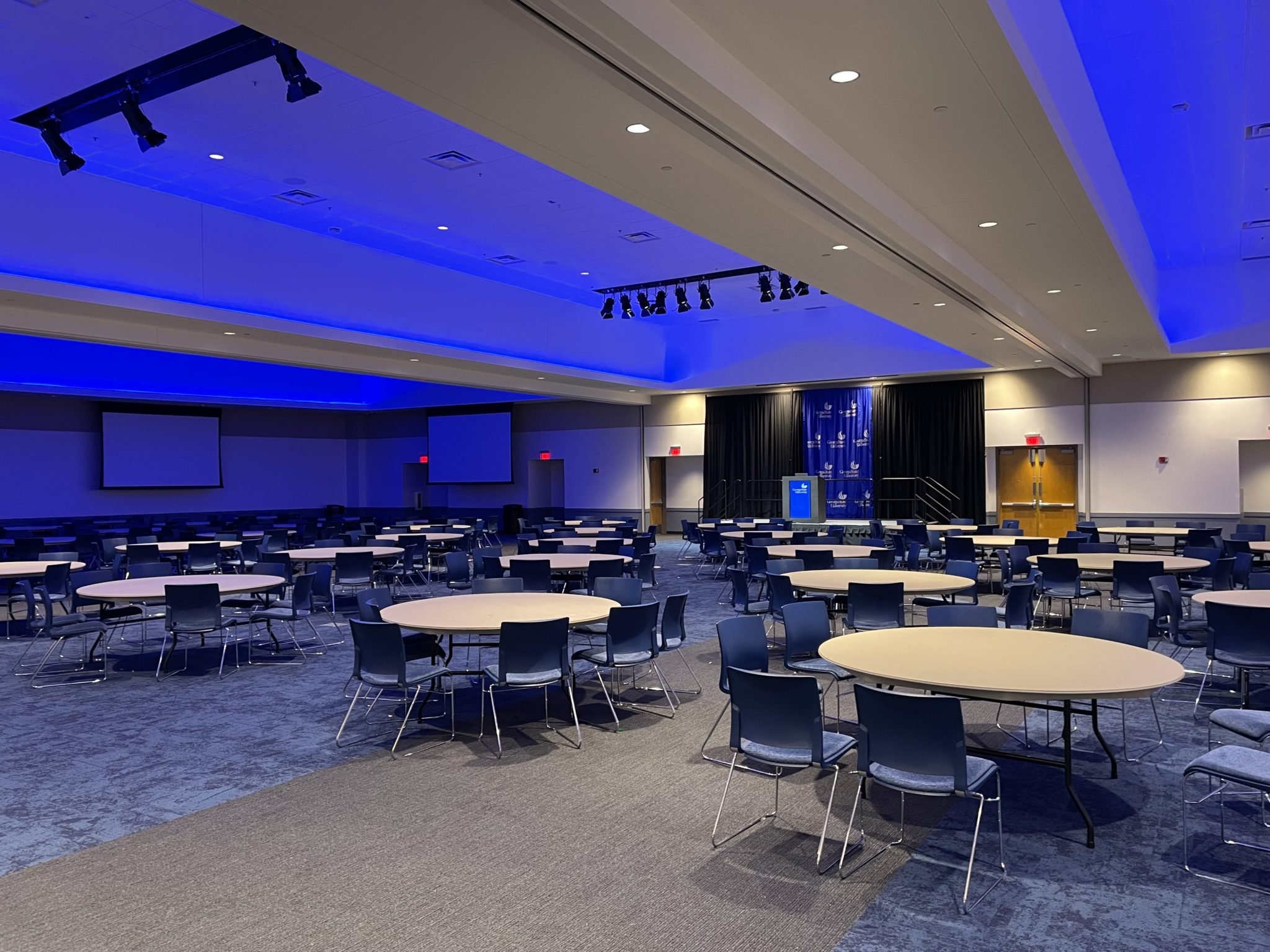 An empty conference or banquet hall with round tables and chairs arranged neatly. The ceiling has built-in lighting and a section of the ceiling is lit with blue LED lights. There is a stage area with curtains and a display screen, with additional screens on the sides of the room.