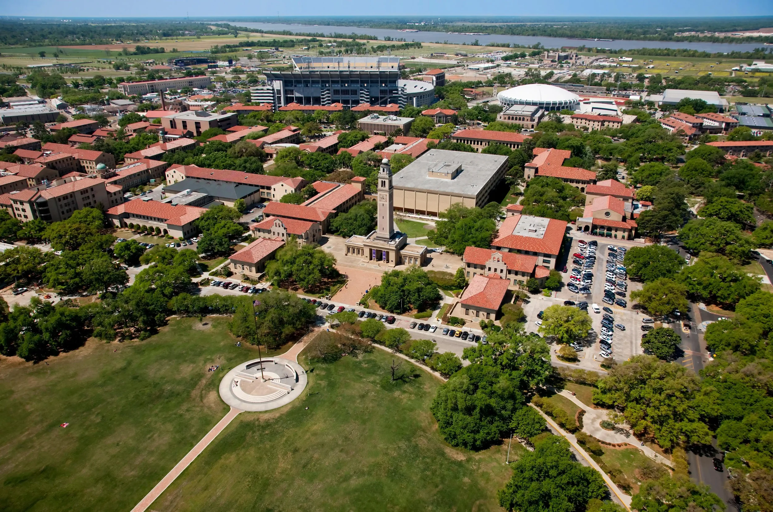 An aerial view of a university campus with red-roofed buildings, a clock tower, a green park with a statue, and sports stadiums, with a river in the background.