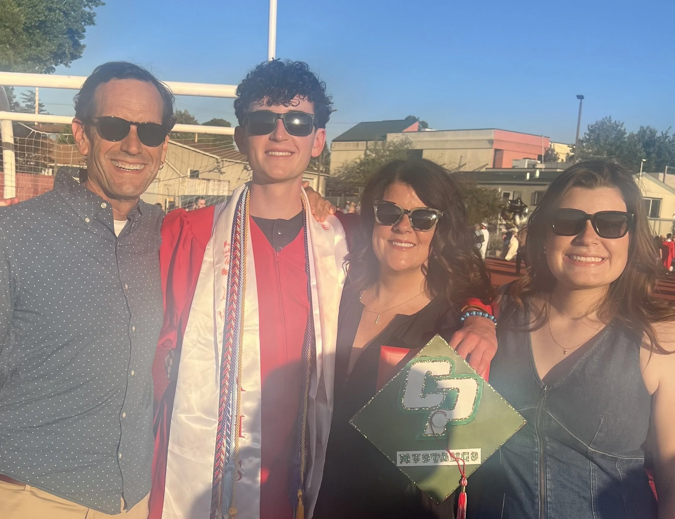 Group of four people smiling, some wearing sunglasses, celebrating graduation outdoors with a clear blue sky.