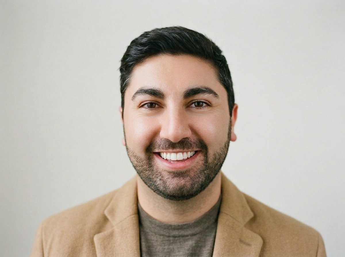 A young man with dark hair, a beard, and a smile, wearing a tan blazer over a gray shirt, standing against a plain light background.